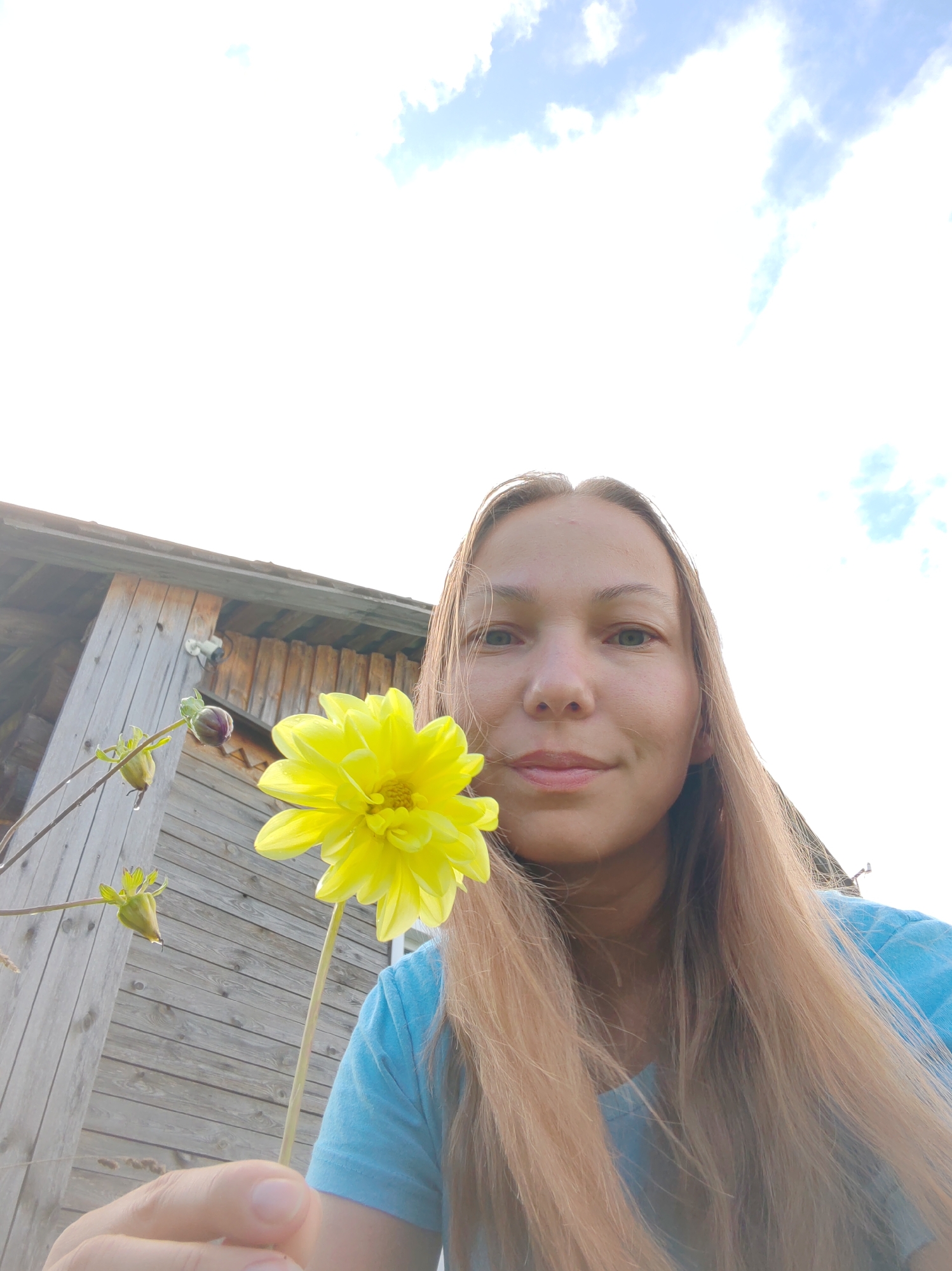 A medium shot, outdoor, low-angle selfie is taken in what appears to be a rural setting. A young woman with long, blonde hair and green eyes is holding a bright yellow flower in her left hand, presenting it towards the camera. She is wearing a blue t-shirt. Behind her, a rustic wooden building with a sloping roof is visible. The sky above is bright and appears to have scattered white clouds. The scene has the feel of a sunny day. No specific location in the city of Чухлэм, Russia can be identified from the visual information.