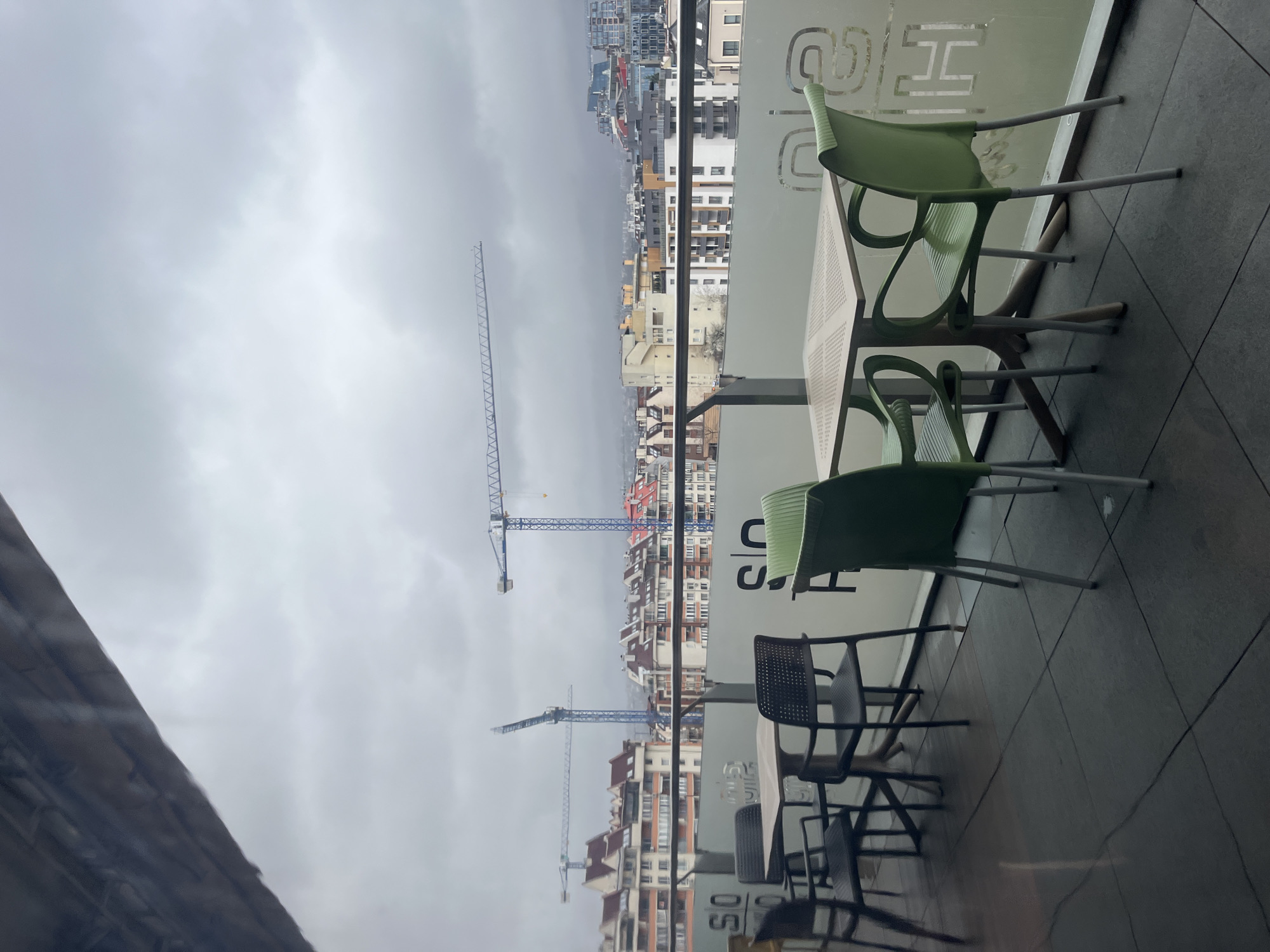 A view from inside a building overlooks a cityscape on an overcast day. Three cranes are visible in the middle distance. In the foreground, there are several tables and chairs, with a mix of green and dark gray chairs. Reflections are visible on the glass, obscuring some of the view. The location is identified as Sofia, Bulgaria based on the architecture and cityscape.