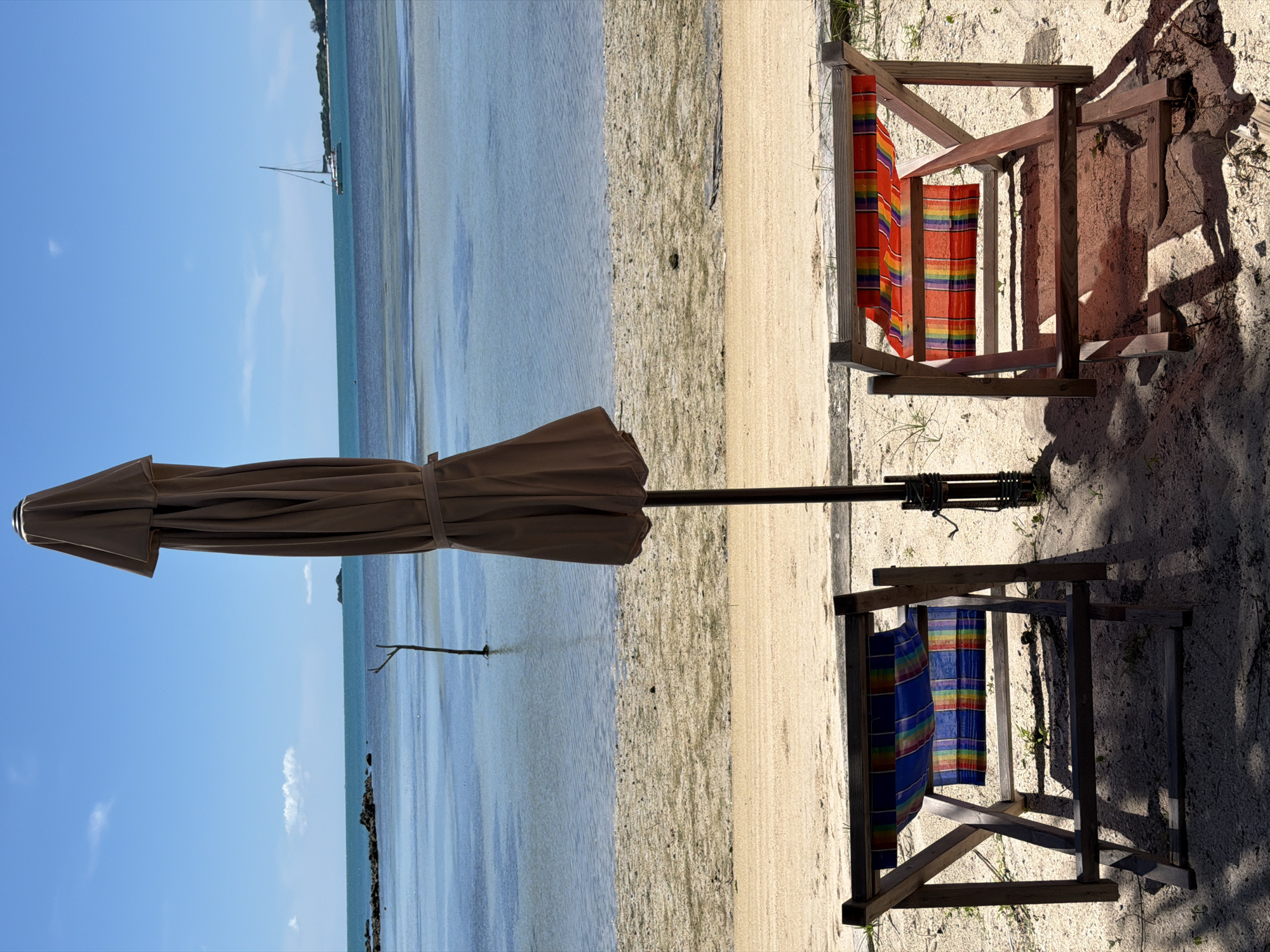 In Baan Thong Krut, Thailand, a serene beach scene unfolds under a bright blue sky with scattered clouds. Two colorful striped deck chairs, one with blue and the other with red and orange stripes, sit on the sandy shore, facing the calm turquoise water. A large, closed beige umbrella stands between them, casting a shadow on the sand. In the distance, a sailboat glides across the ocean, and a small island is visible on the horizon. The shallow water reveals sandbanks and a few scattered rocks, hinting at a low tide. A solitary wooden pole stands in the water near the shore. The scene conveys a peaceful, idyllic beach setting.