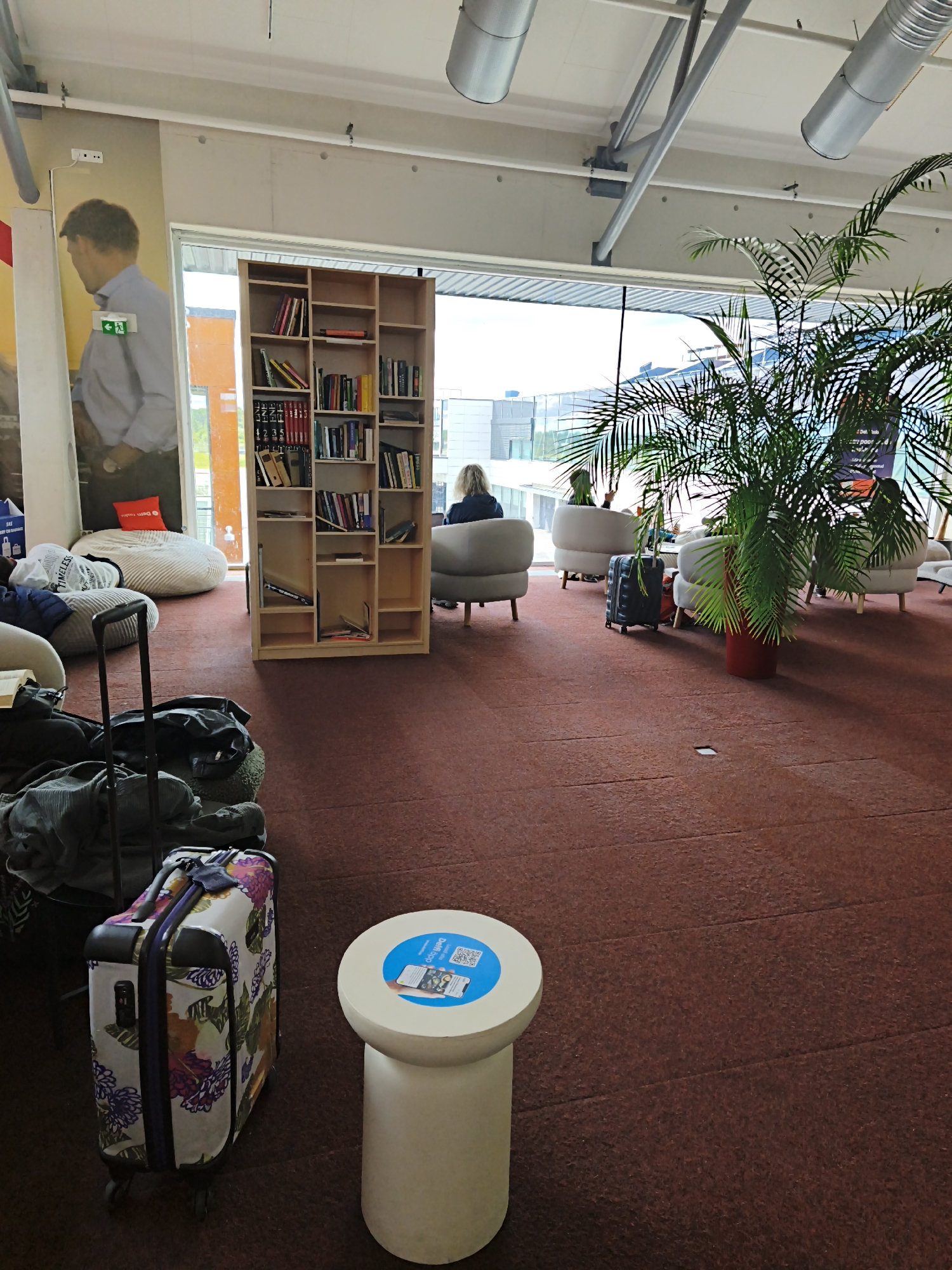 In Tallinn, Estonia, within a modern airport lounge, a spacious room with large windows offers a view of the airport tarmac. The room is furnished with a light wood bookshelf filled with books, several plush lounge chairs, and beanbag chairs. In the foreground, a white cylindrical table with a blue circular advertisement featuring a QR code is positioned next to a floral-patterned suitcase. Several other suitcases and bags are scattered around the seating area. A large potted palm tree stands near the window, adding a touch of greenery to the space. One person is seated in a lounge chair facing the window, their back to the camera, while a few other individuals are visible in the background, also seated. The overall atmosphere appears to be one of relaxed waiting.