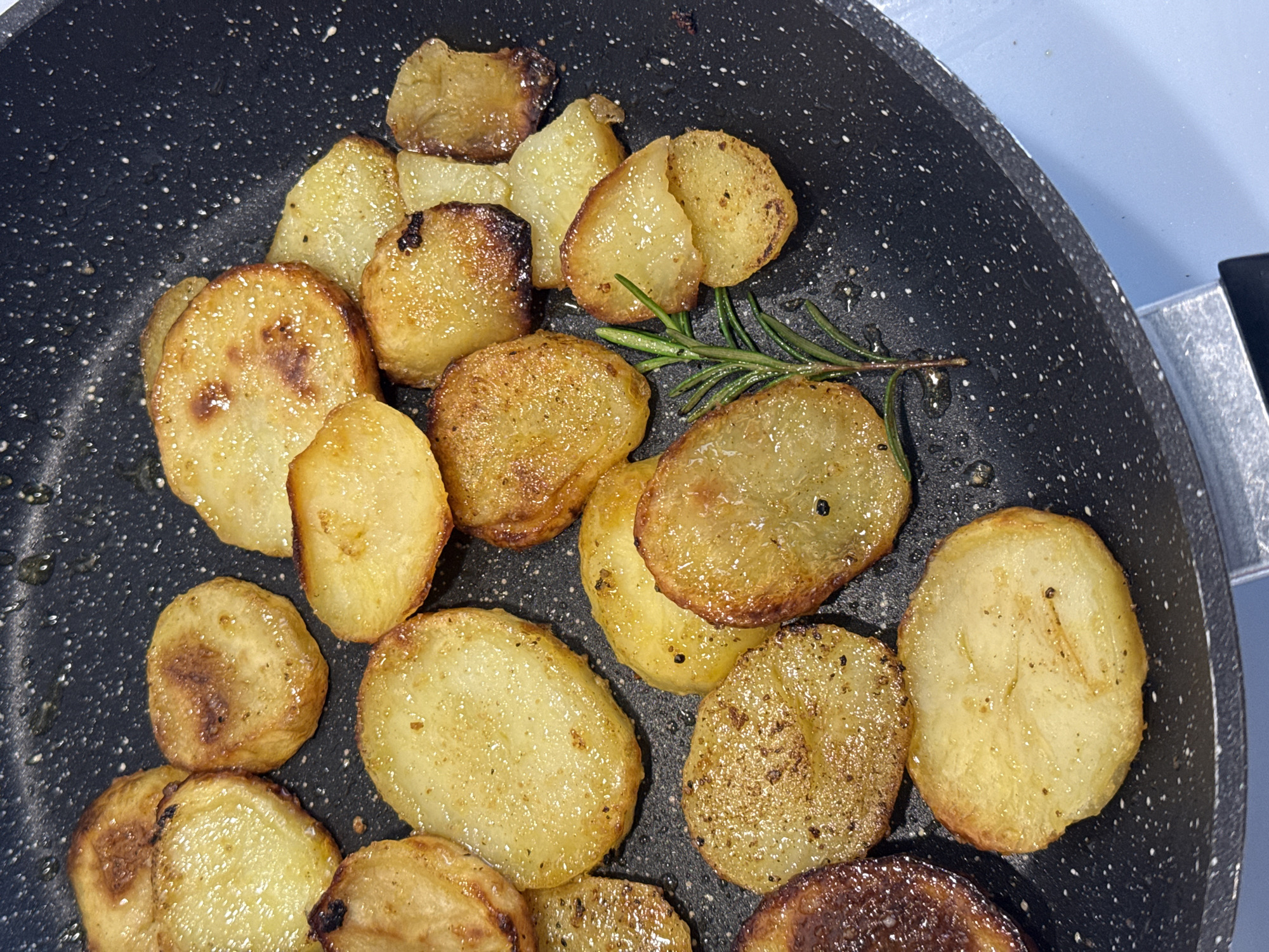 An overhead shot captures a close-up of thinly sliced potatoes frying in a black, speckled non-stick pan. The circular potato slices are primarily golden-brown and yellowish, exhibiting varying degrees of caramelization and crispiness, with some darker browned edges. Small, dark specks, likely seasoning like pepper or herbs, are visible on many of the potato surfaces.
Amidst the potatoes, a fresh sprig of rosemary with green needles is placed, adding a contrasting touch of color and suggesting the flavoring for the dish. Traces of oil or fat are visible as shiny areas in the pan, reflecting the light. The pan's surface shows small, white speckles, characteristic of some non-stick coatings.
In the upper right and along the right edge of the frame, a light blue surface is partially visible, suggesting a countertop or adjacent kitchen appliance. The image depicts the activity of cooking, specifically pan-frying potatoes. There are no people visible, and no discernible text or specific time of day. The scene is an indoor kitchen setting, with the context provided as Milan, Italy, although no specific Italian cues are present in the image itself.