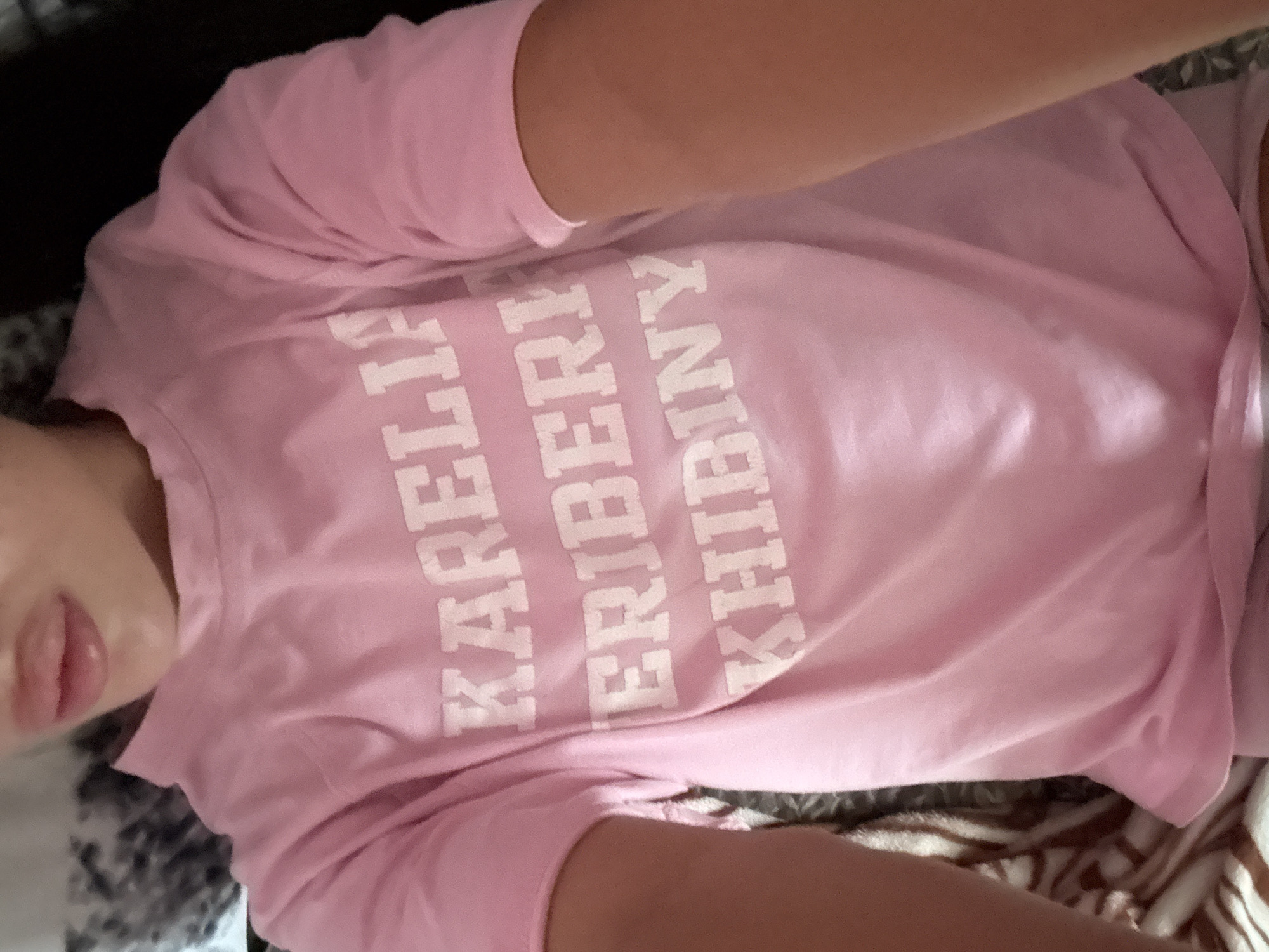 A person is wearing a pink t-shirt with white text on it, which reads "KARELIA ERIBERI KHIBINY." The person's lips are visible at the top of the frame, and their arms are to the sides. The background is blurred and appears to be a bed with a patterned blanket. The overall scene suggests a casual, indoor setting. There is no information about the city of Basarabeasca, Moldova in the image.