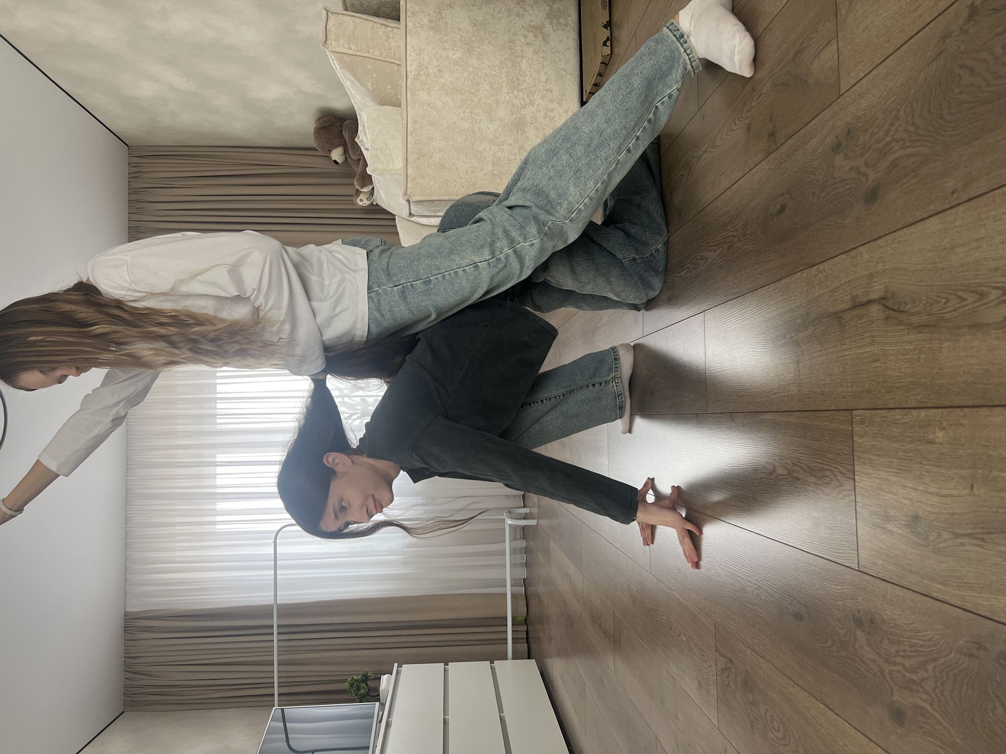 Two women are engaged in a playful activity indoors. One woman, wearing a black top and light blue jeans, is positioned on her hands and knees. The second woman, wearing a white top and light blue jeans, is sitting on the back of the first woman. The scene takes place on a wooden floor, with furniture including a light-colored couch, white drawers, and a television visible in the background. The room has light walls and a window covered by white blinds and light brown curtains. There is no indication that the image was taken in Votkinsk, Russia.