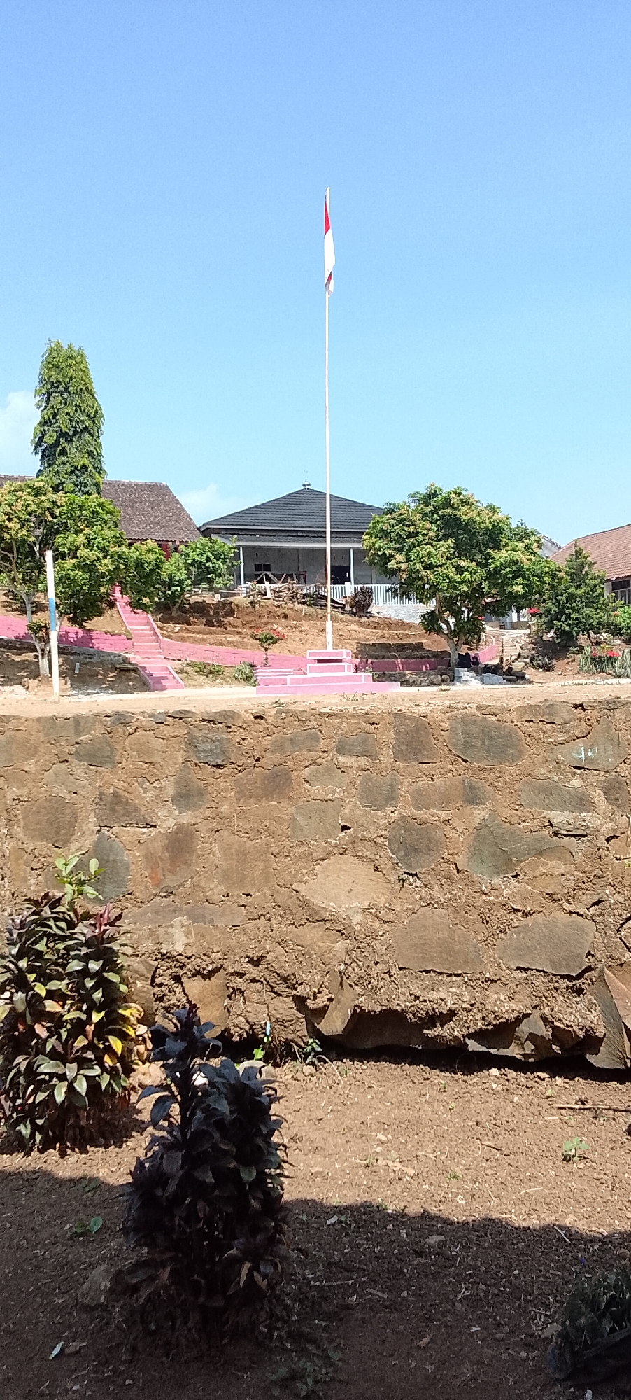 This image captures an outdoor scene in Jepara, Indonesia, under a clear blue sky. In the center, a flagpole stands tall, adorned with the Indonesian flag, white and red. The flagpole is positioned on a tiered platform, bordered by pink accents. To the left, a set of pink stairs ascends a slope, leading towards buildings in the background. These buildings feature traditional Indonesian architecture with dark tiled roofs and open verandas. Lush green trees are interspersed throughout the landscape, providing shade and color. In the foreground, a rough stone wall forms a natural barrier. In front of the wall, several plants with dark green and reddish leaves are visible, adding to the natural beauty of the setting. The ground is covered with soil and a scattering of small plants. The overall atmosphere suggests a serene and well-maintained outdoor space, possibly a school or a public area in Jepara.