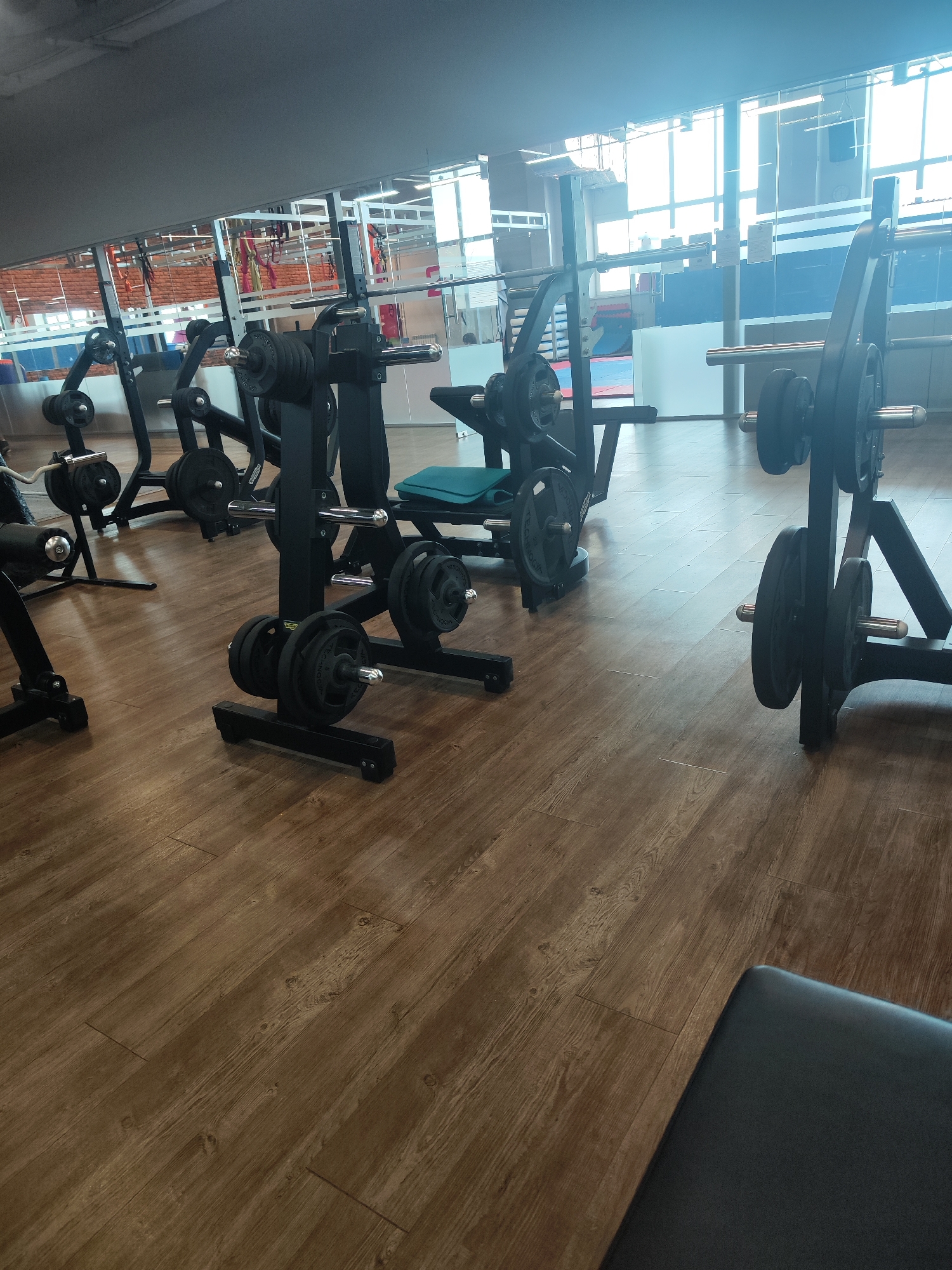 The image shows the interior of a fitness center in Chaikovsky, Russia. Multiple weight training machines are arranged on a wooden floor. The machines are black and loaded with weight plates. In the background, there are windows, a white wall, and a partial view of the interior through the windows. The lighting is bright, suggesting natural light from the windows. No people are visible.