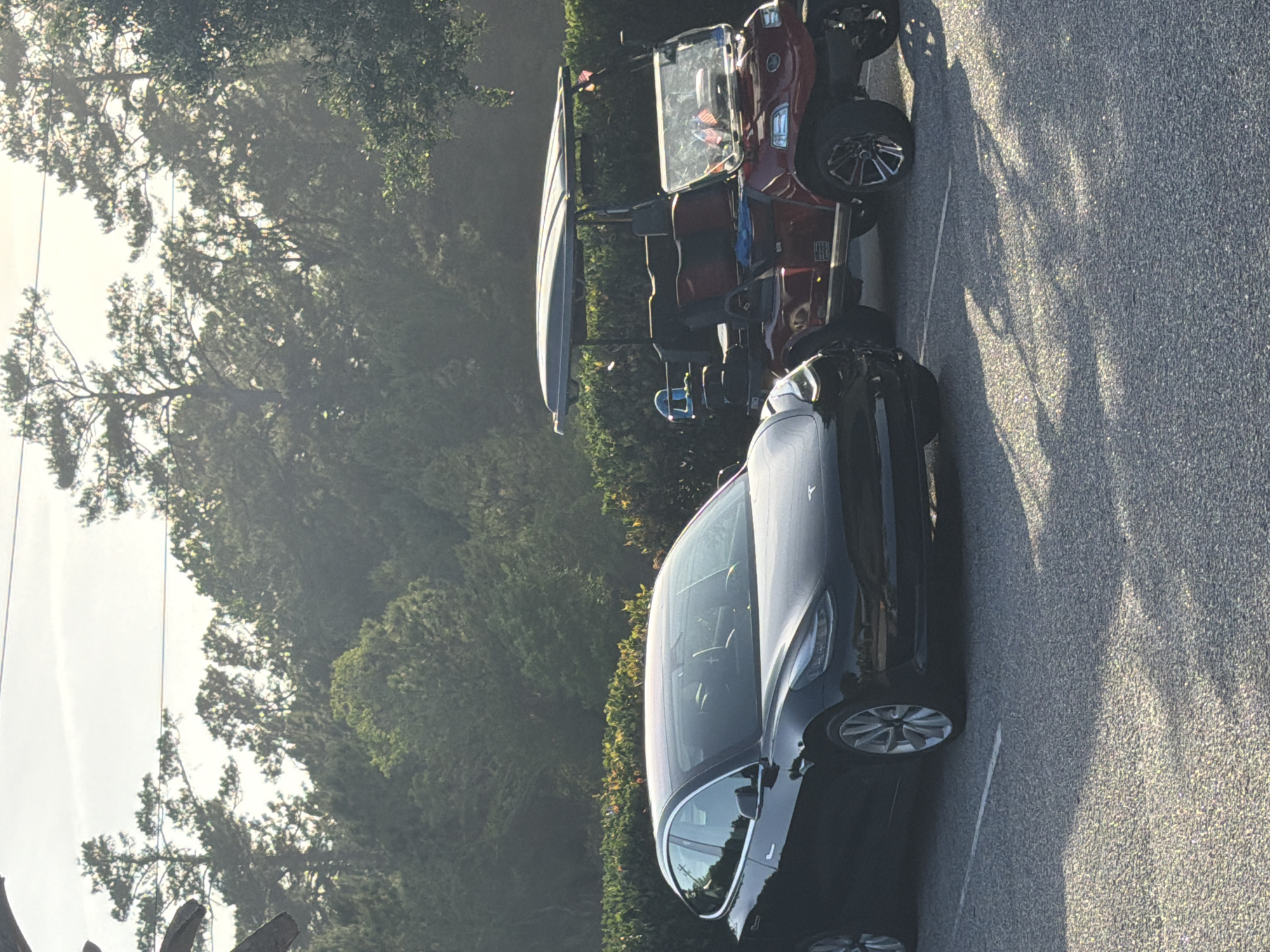 A parking lot with a black sedan and a red golf cart in Myrtle Beach, United States of America. The car is parked next to the golf cart. The vehicles are on an asphalt surface, with trees visible in the background. The lighting suggests it is daytime.