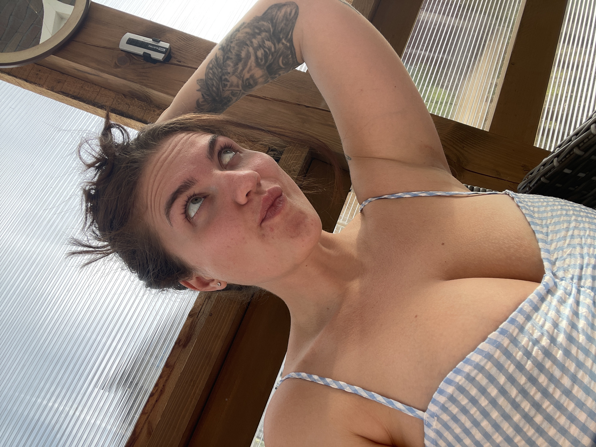 The image shows a woman with an arm raised over her head, revealing a tattoo on her upper arm. She is wearing a light blue and white checkered dress with thin straps. The background shows what appears to be a partially covered outdoor structure with translucent panels. The location cannot be determined from the image alone; the reference to "Мосметаллоконструкция Онт, Russia" does not provide identifiable landmarks or context within the image.