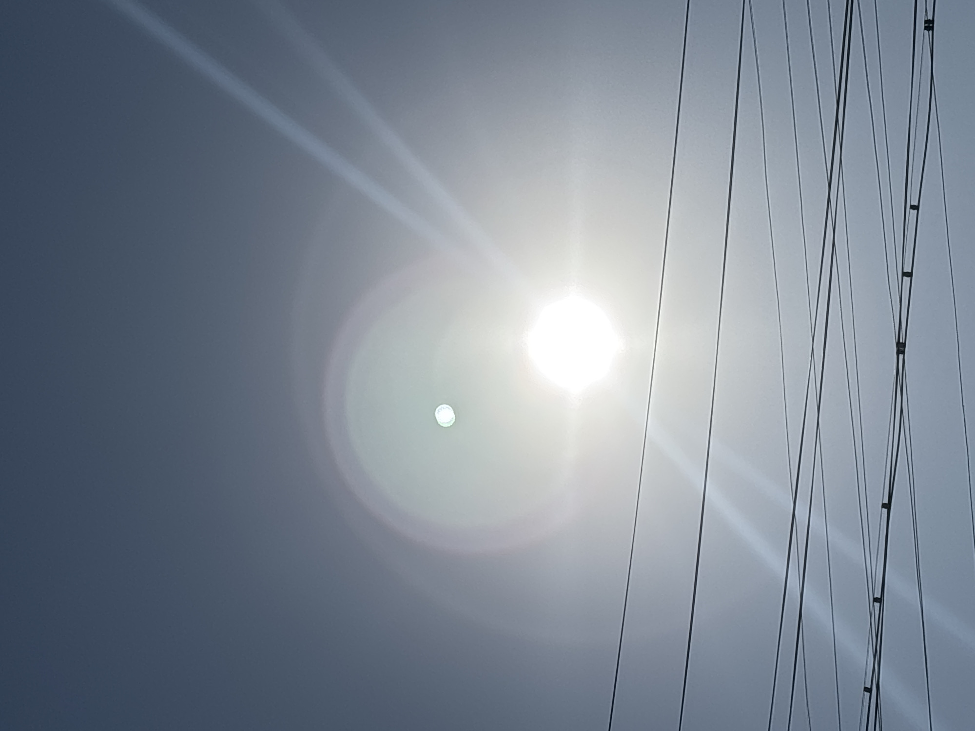 The image shows a clear sky with the sun prominently featured, causing lens flare that creates bright circular patterns and streaks of light. A smaller, circular light source, possibly the moon or another bright object, is visible within the larger sun flare. Several power lines crisscross the lower portion of the frame, casting faint shadows on the sky. The overall scene is devoid of human figures, buildings, or any specific landmarks that would pinpoint a location within Taraz city administration, Kazakhstan. The environment is a sky with overhead wires, suggesting an outdoor urban or suburban setting.