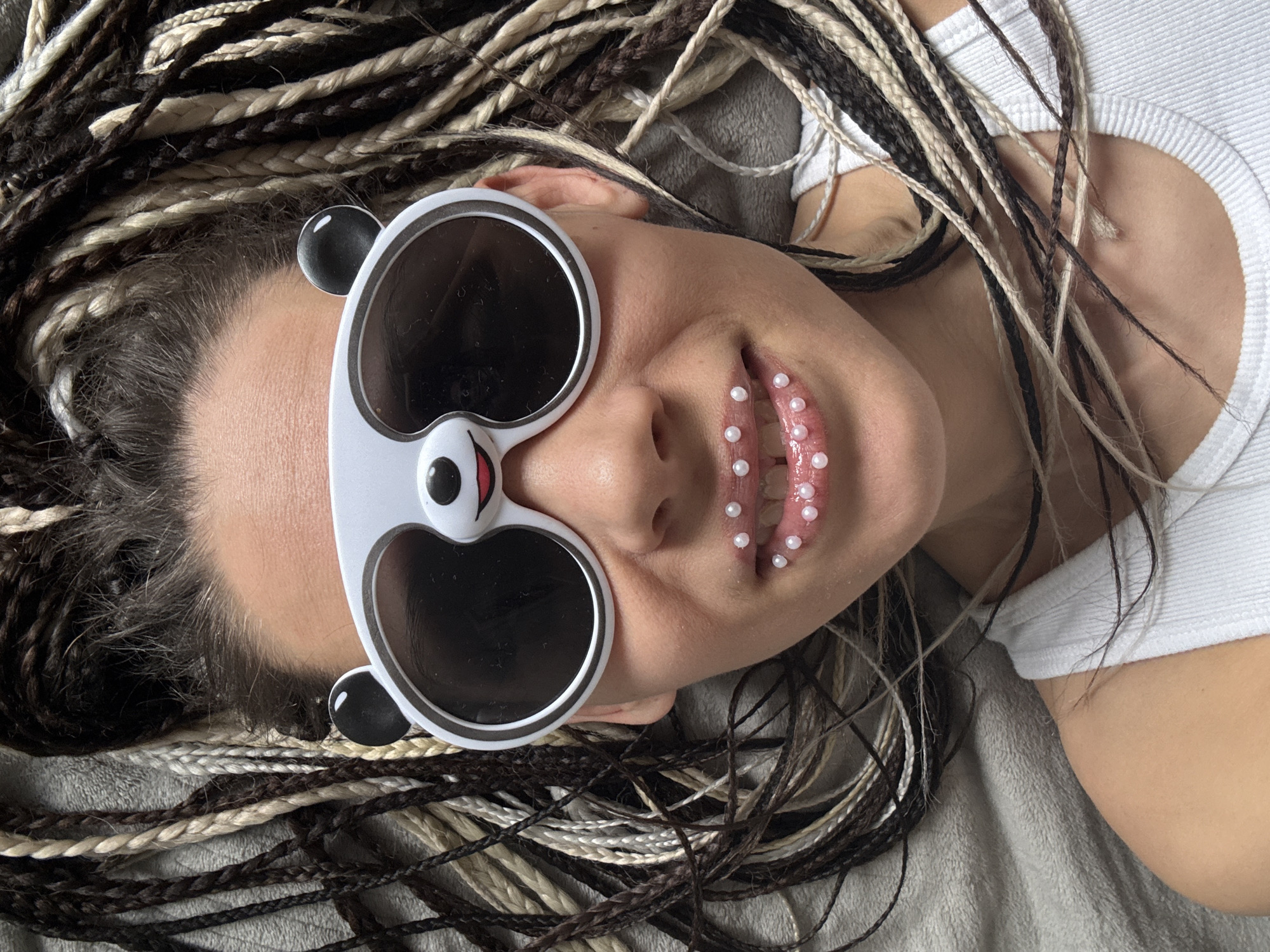 An overhead close-up shot captures a person lying down, showcasing their unique style and accessories. The subject, likely female, is seen from the chest up, with their head angled slightly to the left. They sport long, multi-toned dreadlocks or braids, a mix of dark brown and light blonde strands, fanned out around their head.
A prominent feature is the white, panda-shaped sunglasses worn over their eyes, complete with black lenses and small black ear details. Their lips are adorned with numerous small, iridescent white pearls or beads, creating an avant-garde or decorative effect. The mouth is slightly agape, emphasizing the lip embellishments.
The person is wearing a simple white ribbed top, the fabric of which is visible around the shoulders and neck. The background consists of a soft, grey, textured fabric, possibly a blanket or cushion, suggesting an indoor setting. The overall mood conveyed is artistic and playful. There is no visible text in the image. This scene takes place in Rusanovo, Russia.