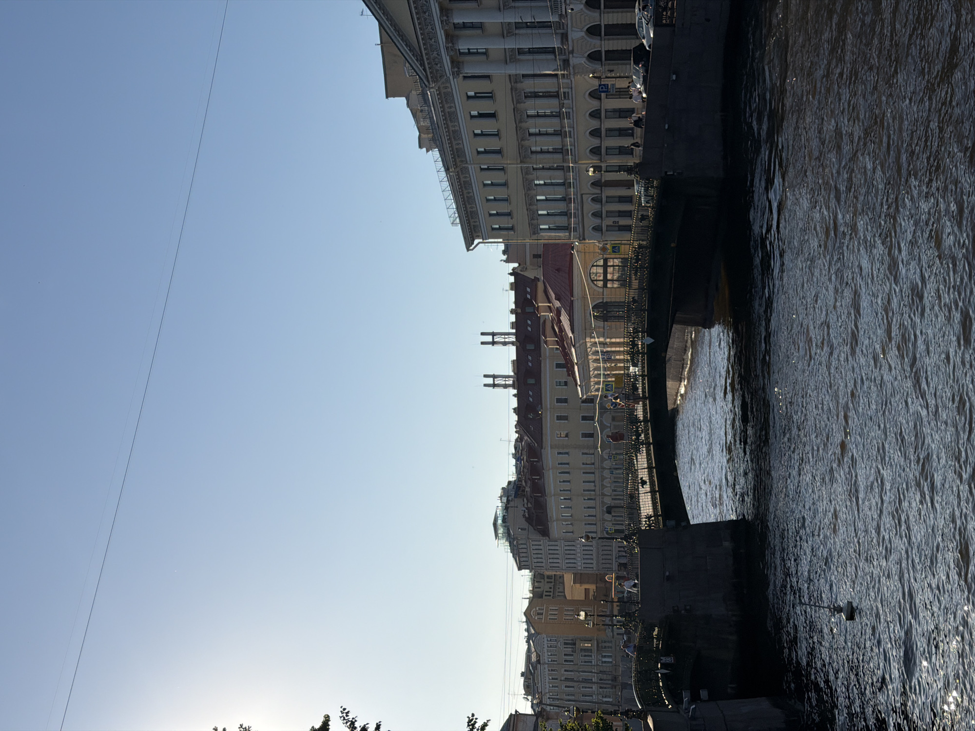 The image shows a canal in Saint Petersburg, Russia, on a sunny day. A pedestrian bridge arches over the dark, rippling water, with people walking across it. The canal is bordered by buildings with classical architecture, featuring rows of windows and ornate facades. The sky is clear and blue, with a single power line stretching across it. The sunlight casts a glint on the water's surface, and the buildings on either side of the canal are illuminated, creating a sense of depth and perspective. The scene depicts a peaceful urban landscape, characteristic of Saint Petersburg's historic districts.