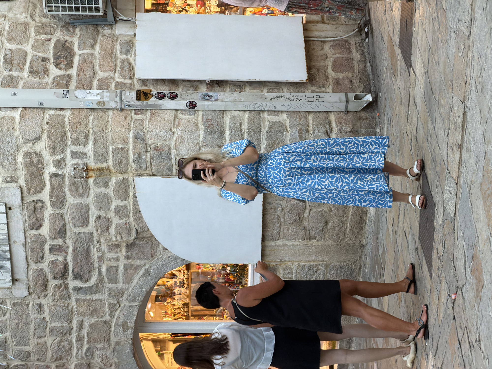 The image captures a street scene in Budva, Montenegro, featuring three women. The architecture suggests an older part of the city, with a weathered stone building forming the backdrop. The woman in the foreground, wearing a blue and white patterned dress, is actively taking a photo with her smartphone. To her left, another woman in a black dress is looking towards a shop window, possibly holding a small item in her hands. A third woman, further to the left and partially visible, wears a white blouse and black skirt, also facing the shop. The shop window displays a variety of items, possibly souvenirs or crafts, illuminated from within. The ground is paved with cobblestones, and a metal drainpipe is visible on the stone wall. The overall atmosphere appears to be a typical European street scene, likely during daytime.