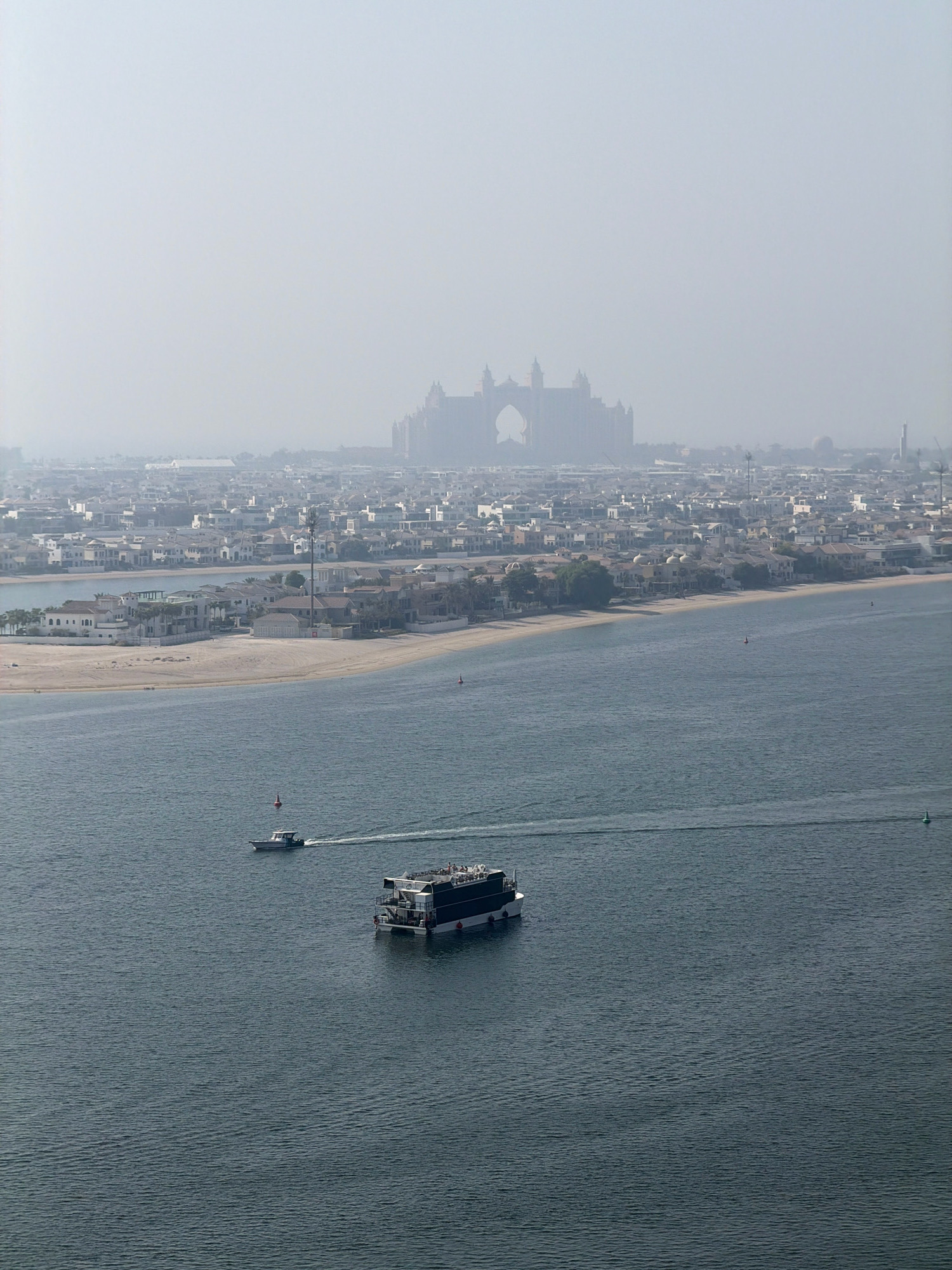 This is a high-angle, wide shot of a hazy cityscape in Dubai, United Arab Emirates, likely taken from a considerable height. In the foreground, a dark-hulled boat with multiple passengers is moving across a blue, rippling body of water. A smaller white boat with at least one person is positioned behind it, also moving.
In the mid-ground, a man-made island with sandy beaches and numerous residential buildings is visible. The architecture of the houses appears to be modern with light-colored walls and dark roofs. A few red buoys are scattered in the water.
The background is dominated by a dense urban sprawl of buildings stretching towards a large, imposing structure in the far distance. This structure, partially obscured by haze, has a distinctive archway and appears to be a resort or hotel. The overall impression is one of a modern, developed coastal city with expansive residential areas and a prominent, large-scale building in the distance. The haze suggests a warm, dry climate.