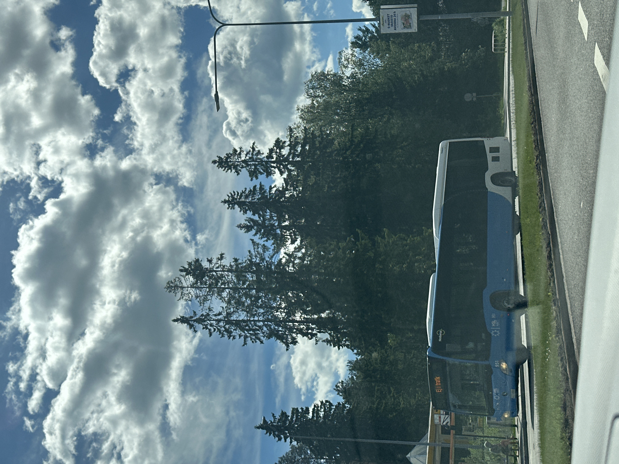 A blue and white bus is driving on a road with a grassy median. The bus has the text "Ej i trafik" on its front. Behind the bus is a line of tall evergreen trees. In the background, a street light with a sign attached is visible. The sky is blue with large, fluffy white clouds. There are some buildings and a yellow bus stop shelter on the left side of the image.