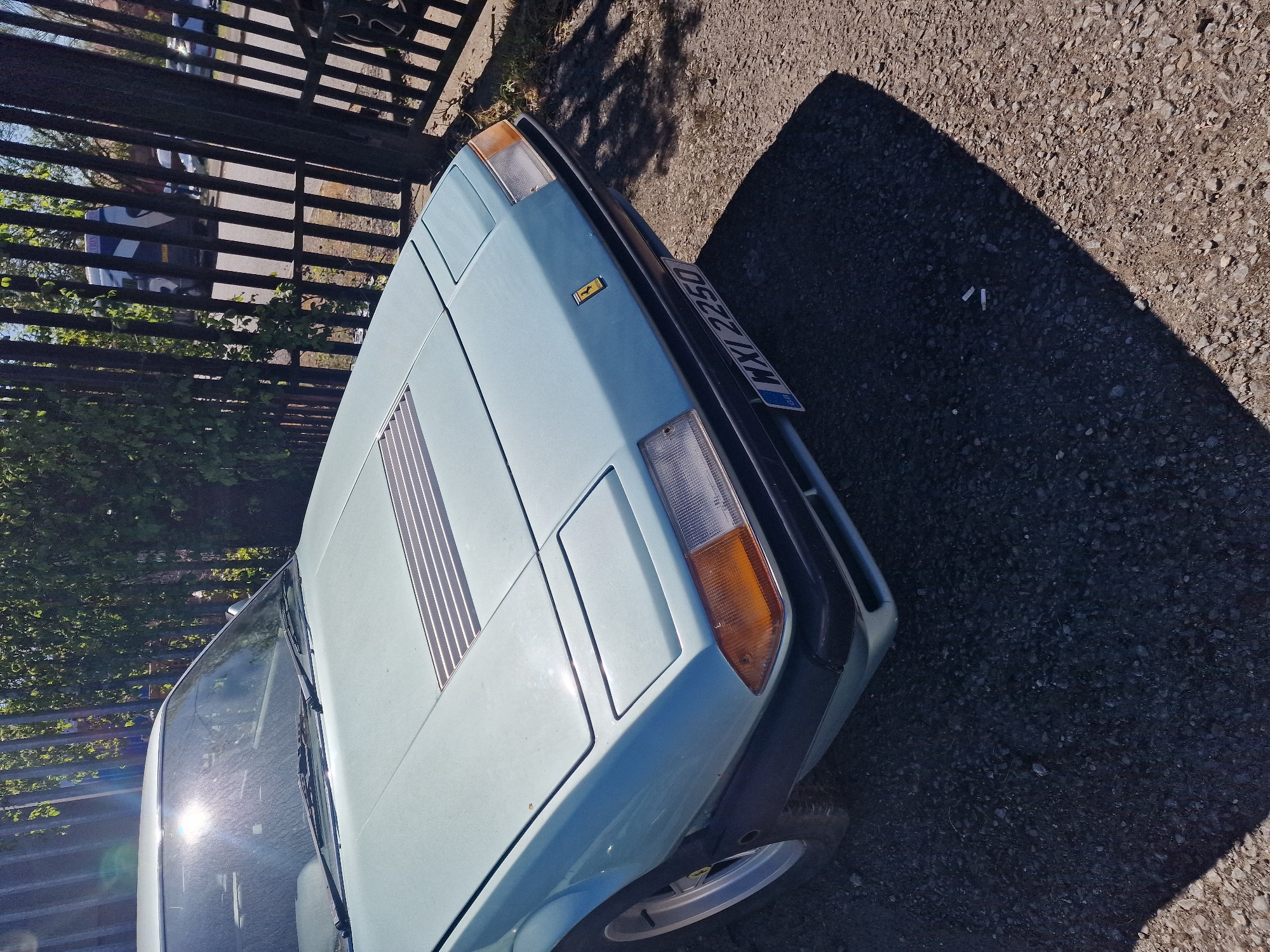 The media file displays a light blue classic Ferrari sports car, likely a 308 GT4, parked outdoors on a gravelly surface. The car is angled, showing its front-right side, including a pop-up headlight cover, an orange turn signal/side marker, and a visible portion of the front grille. A distinctive yellow rectangular Ferrari emblem is on the front hood, which also features narrow, horizontal vents or louvers. The car's windshield shows significant sun glare and reflections.
To the left of the car, a dark metal fence runs horizontally across the frame, with green foliage visible behind it. Strong, dark shadows are cast on the gravel in the foreground and to the right, indicating bright sunlight. The time of day appears to be mid-day or early afternoon due to the harsh lighting and distinct shadows. The weather is clear and sunny.
The car bears a UK license plate, clearly displaying "GB" on the left and the registration "WYL 2250." This, combined with the provided context, places the scene in South Tyneside, United Kingdom. There are no people visible, and the car is stationary, suggesting it is simply parked. Two small white cylindrical objects, possibly cigarette butts, are visible on the shadowed ground area in one of the crop images.
