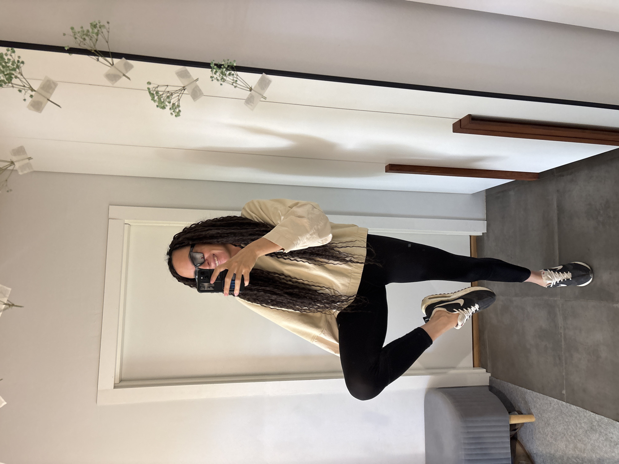 A person is captured in a mirror selfie within an indoor setting in Chaikovsky, Russia. The subject is a woman with long, dark dreadlocks or braids, wearing black-rimmed glasses and smiling slightly. She is dressed in a loose-fitting, light beige or cream-colored long-sleeved top, black leggings, and black athletic shoes with white accents and the Nike logo visible. She holds a black smartphone, taking the picture of her reflection.
The background visible in the mirror shows a contemporary, well-lit interior. The walls are predominantly white, with some light grey sections. Along the top edge of the mirror, small sprigs of light green foliage, possibly baby's breath, are decoratively taped to a wire or string. To the right, two horizontal wooden shelves or decorative ledges extend from the wall. The floor is a dark grey, appearing to be concrete or large tiles. A partial view of a light grey upholstered bench or stool is visible in the lower right corner. The lighting is bright and even, suggesting either good natural light or strong indoor lighting. The overall mood conveyed by the subject's expression is positive.