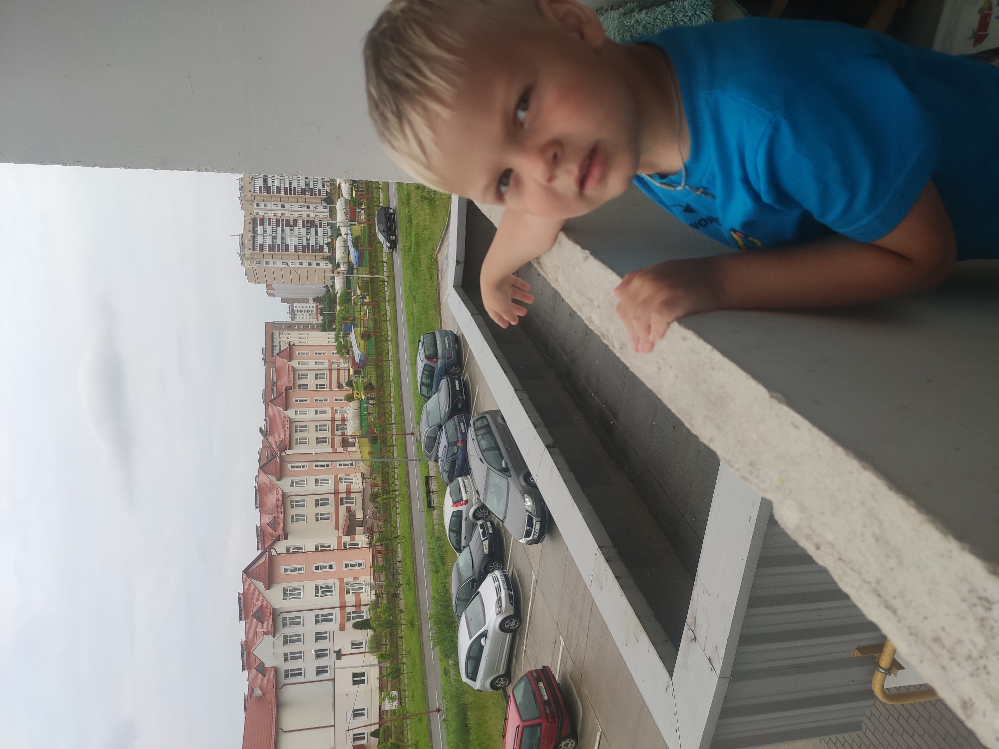 The image shows a young boy with blond hair and blue eyes, wearing a blue t-shirt, leaning on a concrete railing on a balcony. He is looking towards the camera with a neutral expression. In the background, there is a street with several parked cars, including a red station wagon, a silver hatchback, a black sedan, and a dark gray station wagon. Beyond the street, there are several apartment buildings of varying heights and architectural styles, and a cloudy sky. The setting appears to be a residential area in Navapolatsk, Belarus. The boy's arm is resting on the railing, and his other hand is also visible on the railing. The image is taken from a slightly elevated perspective, looking down at the street and the boy.