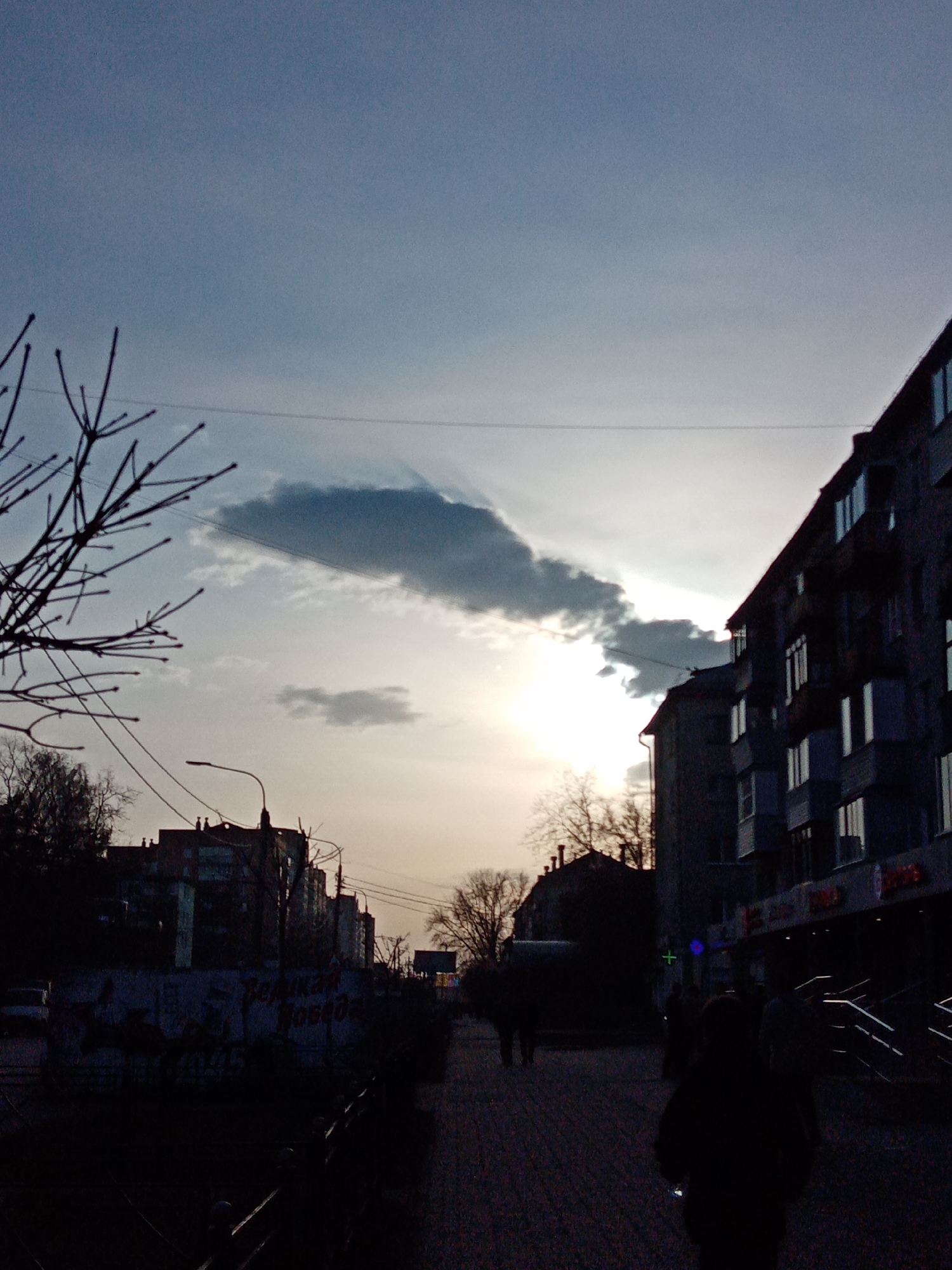 The image presents a dusk scene in Elektrostal, Russia. The sky is a gradient of blue and yellow where the sun sets. There are dark clouds in the sky. Bare tree branches are visible in the foreground on the left side of the image. The lighting is dim, and the city buildings appear as dark silhouettes. There is a building on the right of the image that appears to be a store. People are walking along the sidewalk. A street light is visible on the left, but it is not illuminated. In the midground there is a wall that appears to have graffiti on it.