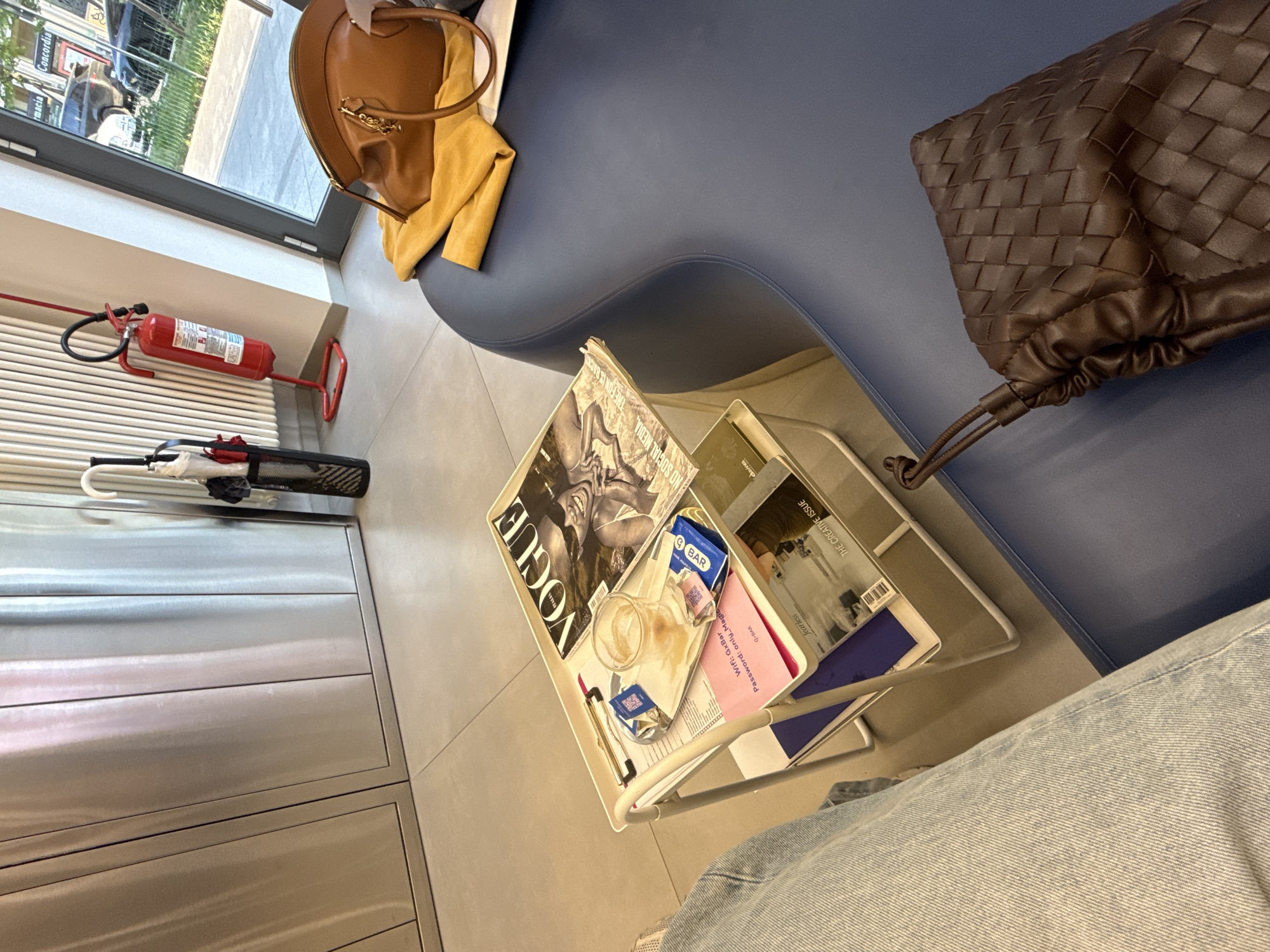 This image depicts a modern indoor waiting area or lounge, possibly within a salon or stylish office, during bright daylight hours. A person's leg, clad in light-wash jeans, is visible in the foreground, suggesting they are seated on a plush, wave-shaped blue sofa.
On the sofa, a tan leather handbag with gold accents rests alongside a yellow garment, while a dark brown woven leather bag is positioned further along the cushion. Adjacent to the seating, a white metal magazine rack or side table holds an assortment of items. Magazines such as "VOGUE ITALIA," "THE CREATIVE ISSUE" by "fearless," and one with the tagline "NO SOCIAL MEDIA, THE FUN IS BACK" are prominently displayed. An empty or half-empty coffee cup, a wrapper, and a small white packet are also inside the rack. A pink card provides details for "Wifi: GxBar" with the "Password: only_Magic" and identifies the location as "Q-BAR." The brand "davines" is also visible on one of the publications.
In the background, against a neutral-toned wall, a red fire extinguisher is mounted, and a stand containing umbrellas is situated beneath it. A large window offers a view of a street scene, including glimpses of buildings and vehicles, with a sign reading "macia Concordia," likely part of a "Farmacia Concordia," a common sight in Milan, Italy. The floor is covered with large, light grey tiles, contributing to the clean and contemporary aesthetic of the space.