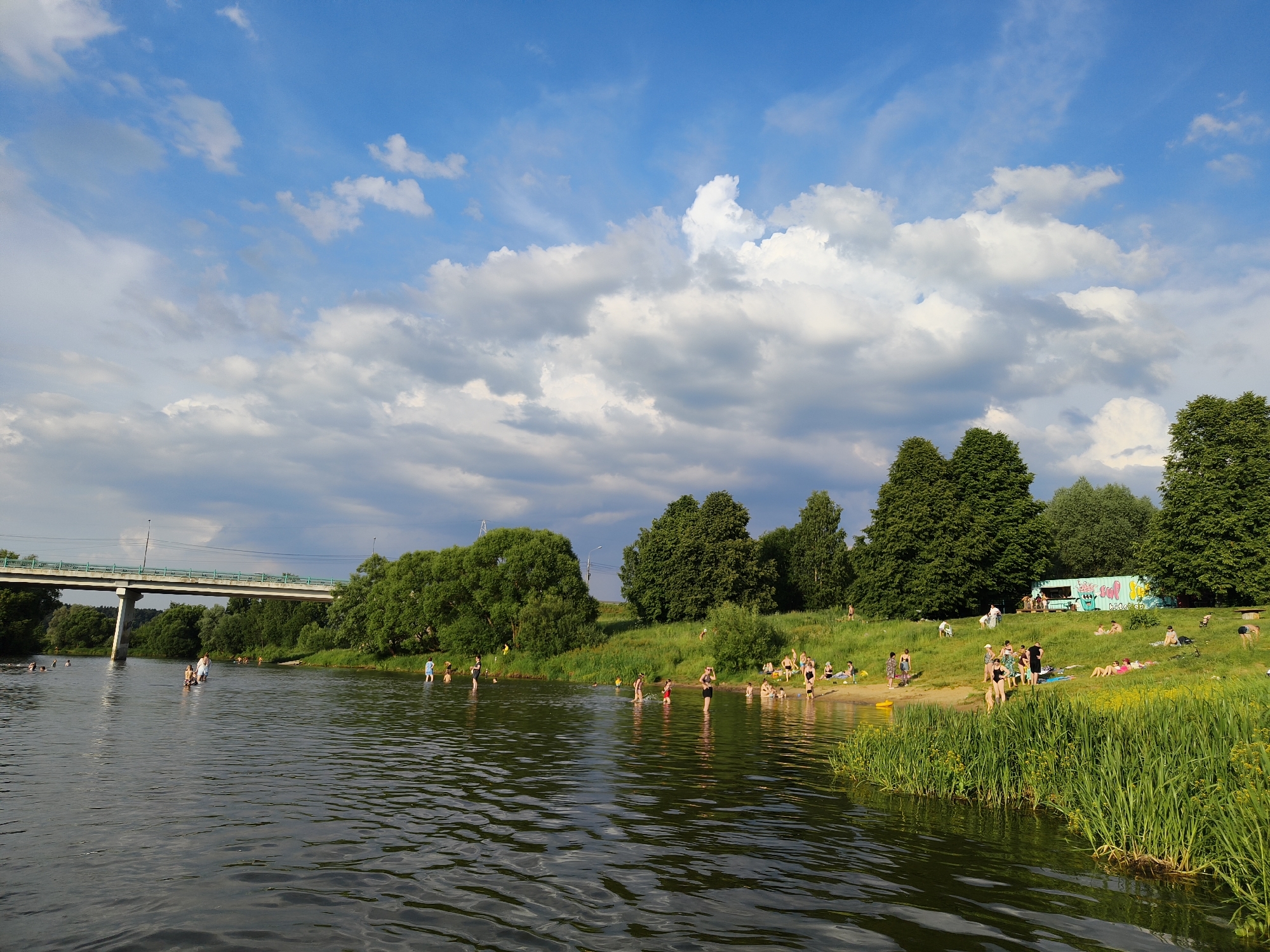The image depicts a sunny day at a riverbank. The river occupies the left and lower portions of the frame, with ripples indicating a gentle current. A bridge spans the river in the background, supported by concrete pillars. Lush green trees line the far bank, and a grassy slope dotted with people is visible on the right bank. Several individuals are in the water, some wading and others swimming. Many people are sunbathing or relaxing on the grassy area and a small sandy beach. A food truck is parked on the grass near a cluster of trees. The sky is a bright blue with scattered white clouds, suggesting fair weather. The scene conveys a relaxed atmosphere of recreation and leisure.