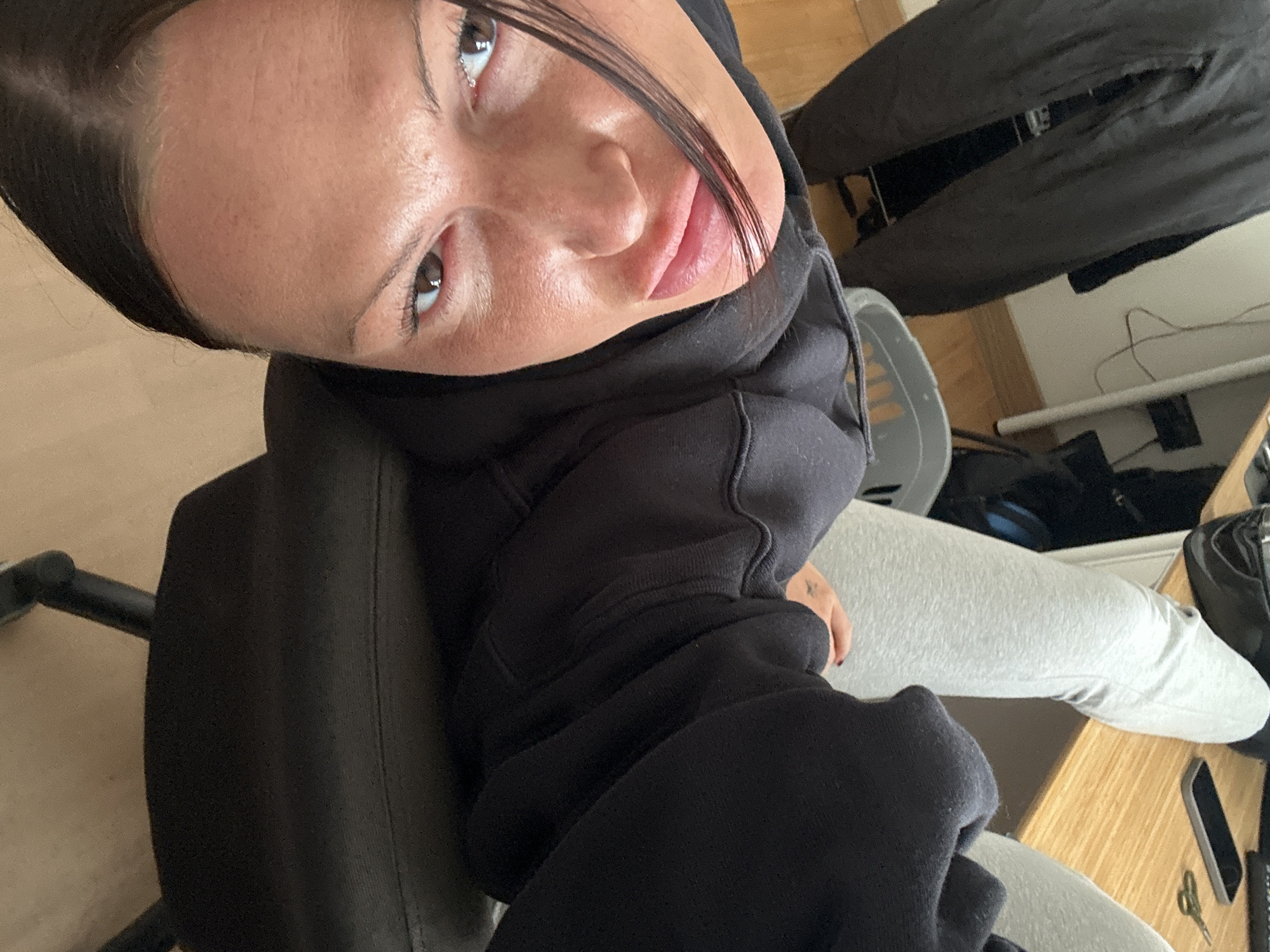 This high-angle, tilted photograph features a young woman as the primary subject, looking upwards towards the camera. She has dark hair, brown eyes, and a neutral, calm expression with a hint of a soft smile on her lips. Her skin shows natural texture.
She is wearing a dark, likely black, hooded sweatshirt or jumper, which covers her upper body and neck. Her lower body, partially visible, is clad in light grey sweatpants or leggings. A small, dark-colored tattoo is visible on her right hand or wrist, and her fingernails are painted with dark polish. She appears to be seated in a dark office chair, parts of which are visible on the left and at the bottom of the frame.
The setting is an indoor space, likely a home office or bedroom in Wealden, United Kingdom. A light-colored wooden desk is visible on the right, holding a black smartphone and a small pair of gold-colored scissors. The background includes a light-colored wooden floor and a plain light-colored wall. A dark jacket or piece of clothing hangs in the distant background, and some cables are visible near the wall. A grey plastic chair or stool is partially seen behind her. The lighting is soft and ambient, suggesting daytime indoors. The overall scene conveys a casual, relaxed moment captured in a personal environment. No visible text is present in the image.