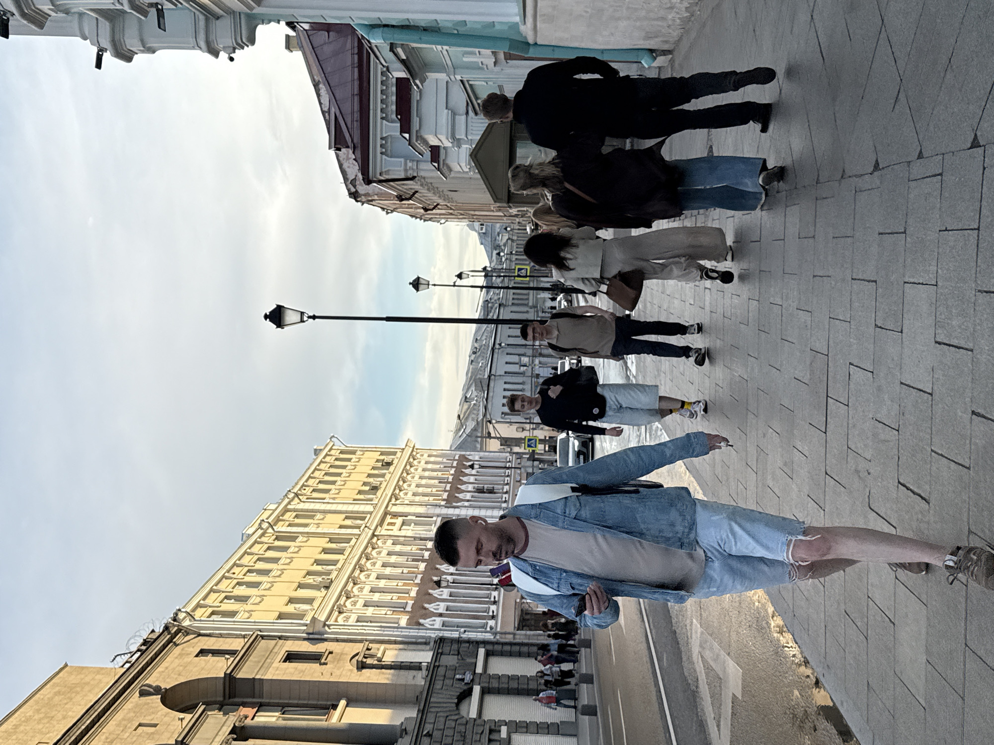 The image depicts a street scene in Moscow, Russia, under a bright, overcast sky, suggesting it's daytime. Several people are walking on a paved sidewalk alongside buildings with classic architectural details. The closest subject is a man in the foreground, wearing a denim jacket, a light-colored t-shirt, and ripped denim shorts. He is looking down at his phone and appears to be holding a cigarette. Further down the street, other individuals are seen walking in both directions. Among them are people wearing casual clothing, including jeans, jackets, and sneakers. The buildings lining the street are multi-story structures with numerous windows, some painted in light colors. A streetlamp is visible in the background, characteristic of an urban setting. The overall atmosphere is that of a typical day in a city. No visible text is present in the image.