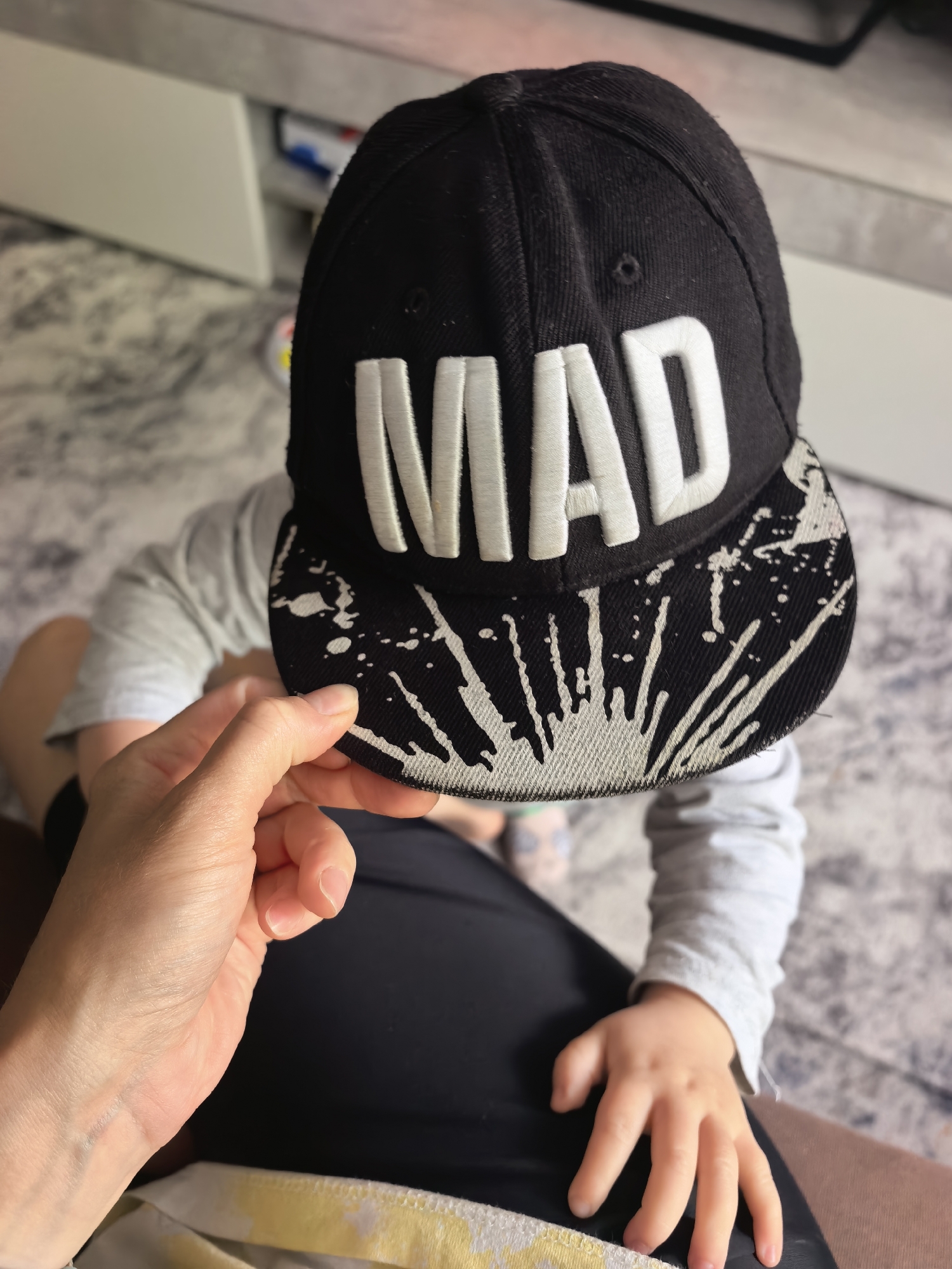 An overhead-angle image depicts an indoor scene involving a child and an adult's hand. The specific location within Sterlitamak, Russia, cannot be visually confirmed from the provided image.
In the foreground, a light-skinned adult hand is visible on the left, gently touching the brim of a black baseball cap worn by a child. The cap features prominent white embroidered letters spelling "MAD" across the front, with the brim displaying a white paint splatter or distressed design. The child's face is completely obscured by the cap. The child is wearing a light grey long-sleeved shirt, with a small, light-skinned hand resting on a dark fabric surface in the lower right, presumably the adult's lap or clothing. The adult is wearing dark-colored lower garments, with a strip of yellow and white patterned fabric visible below. The background consists of a light-colored floor, possibly carpeted or tiled, with a subtle gray and white marbled or textured pattern. Further in the blurred background, a white furniture piece or wall element is partially visible, indicating a domestic indoor setting. The lighting is bright, illuminating the scene clearly.