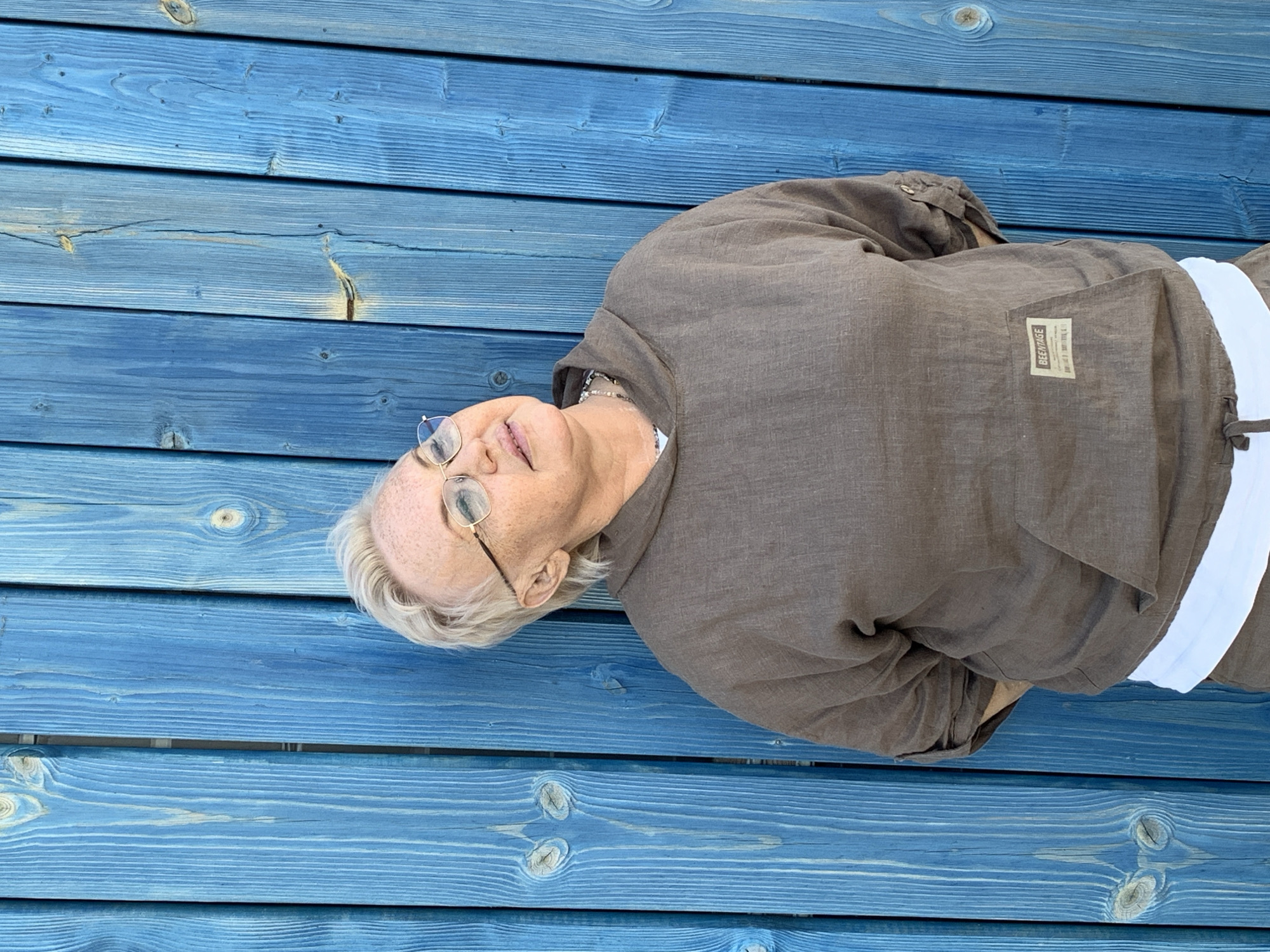 A woman lies on a blue wooden surface. She is wearing glasses and a brown linen top with a white accent around her waist. A label is visible on the top. The location is Chaikovsky, Russia.