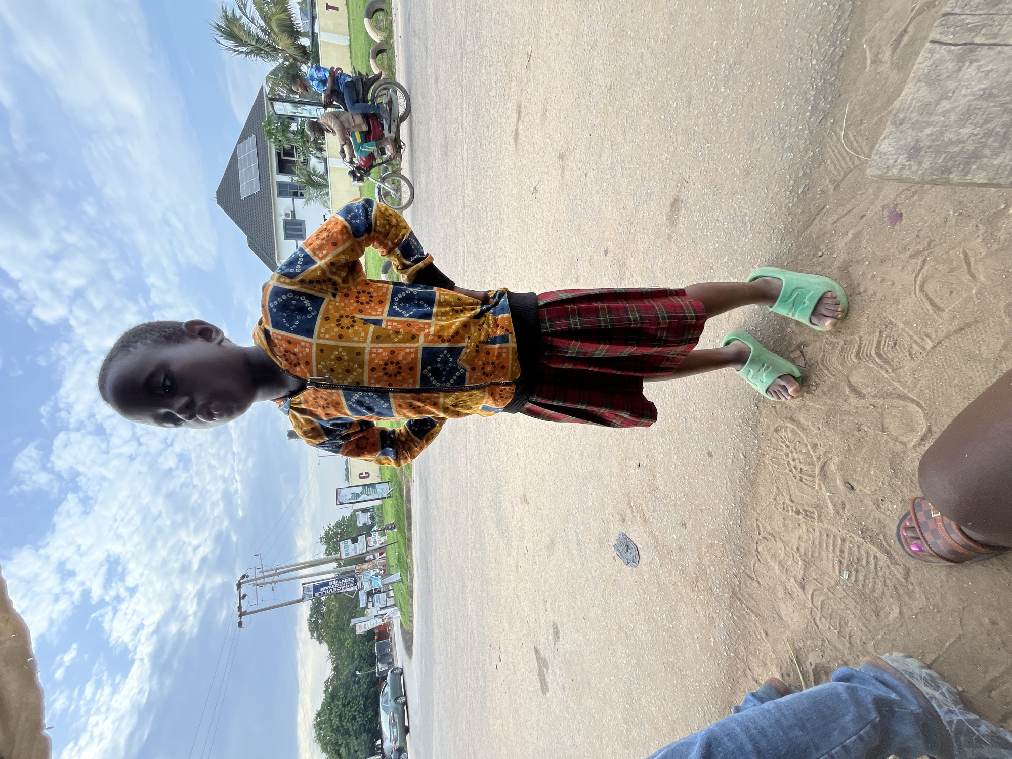 A young Black child stands on a sandy area beside a paved road, facing slightly away from the camera. The child wears a vibrant orange, blue, and yellow patterned zip-up hoodie over a red and black plaid skirt and light green slide sandals. The child's hands are on their hips, and their expression is looking upwards. Behind the child, a white building with solar panels on its roof is visible, along with a motorcycle carrying two people on the road. The environment appears to be a semi-urban setting, possibly on the outskirts of Abuja, Nigeria, characterized by utility poles, street signs, and some greenery. The ground is a mix of sand and pavement, with tire tracks and footprints visible in the sand. The sky is partly cloudy.