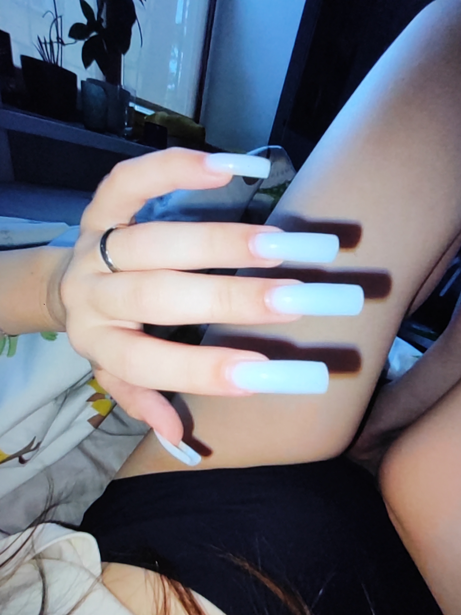 The image shows a close-up of a person's hand with long, light blue acrylic nails, and a portion of their legs. The person is wearing a silver ring on their left ring finger. The background is blurred and appears to be an indoor setting with some furniture and plants visible on a shelf. There is also a hint of a black fabric garment on the lower part of the image. The lighting is dim with a blue hue. There is no discernible information within the image to suggest the location in Fergana City, Uzbekistan.