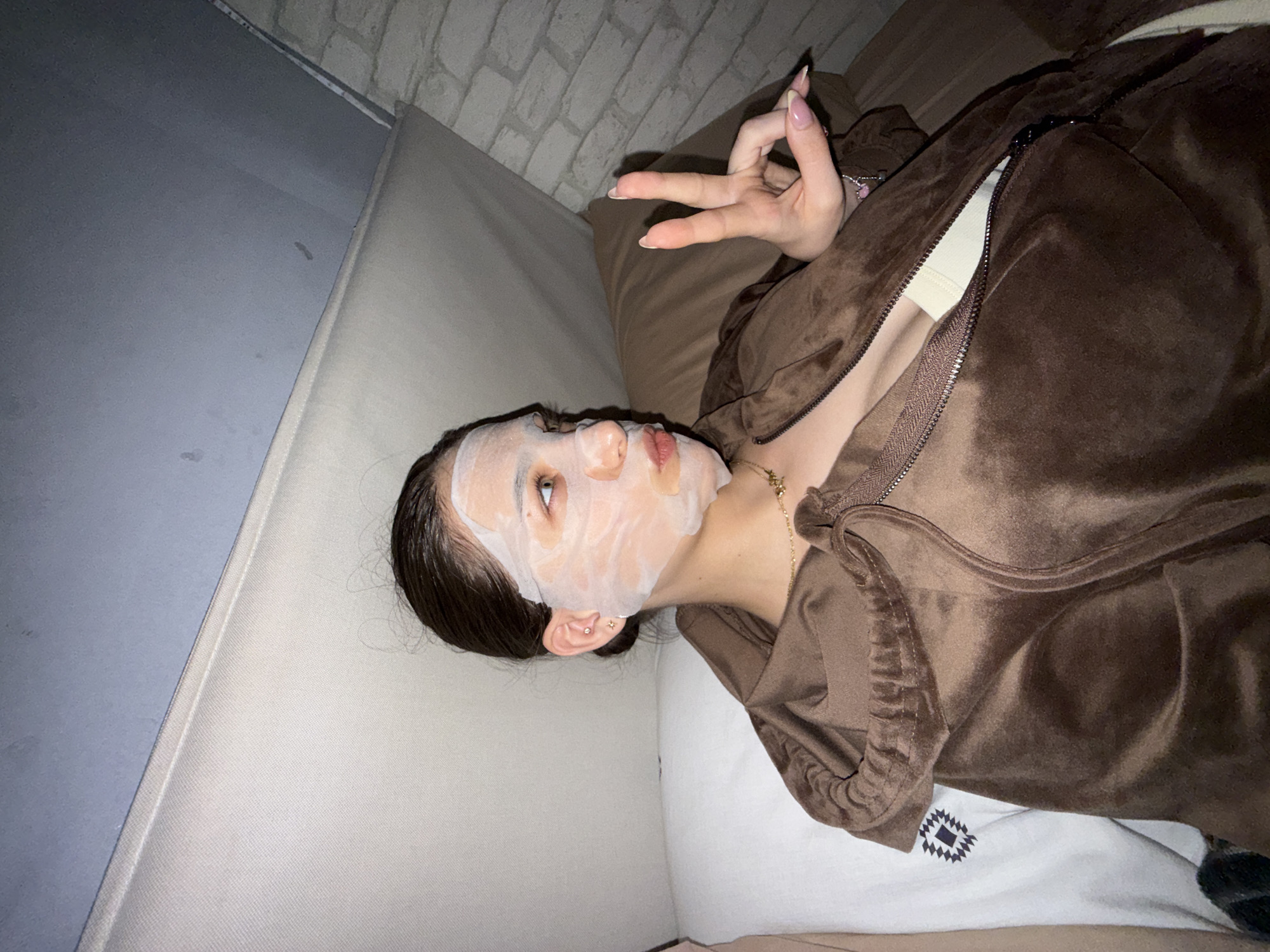 A young adult female lies on a light-colored sofa, facing upward and slightly angled to the right. She wears a white face mask and a brown zip-up hoodie. Her right hand is raised, making a "peace" sign. A light-colored brick wall is visible in the background. The setting appears to be indoors, but there is no identifiable information to indicate a specific location in Sofia, Bulgaria.