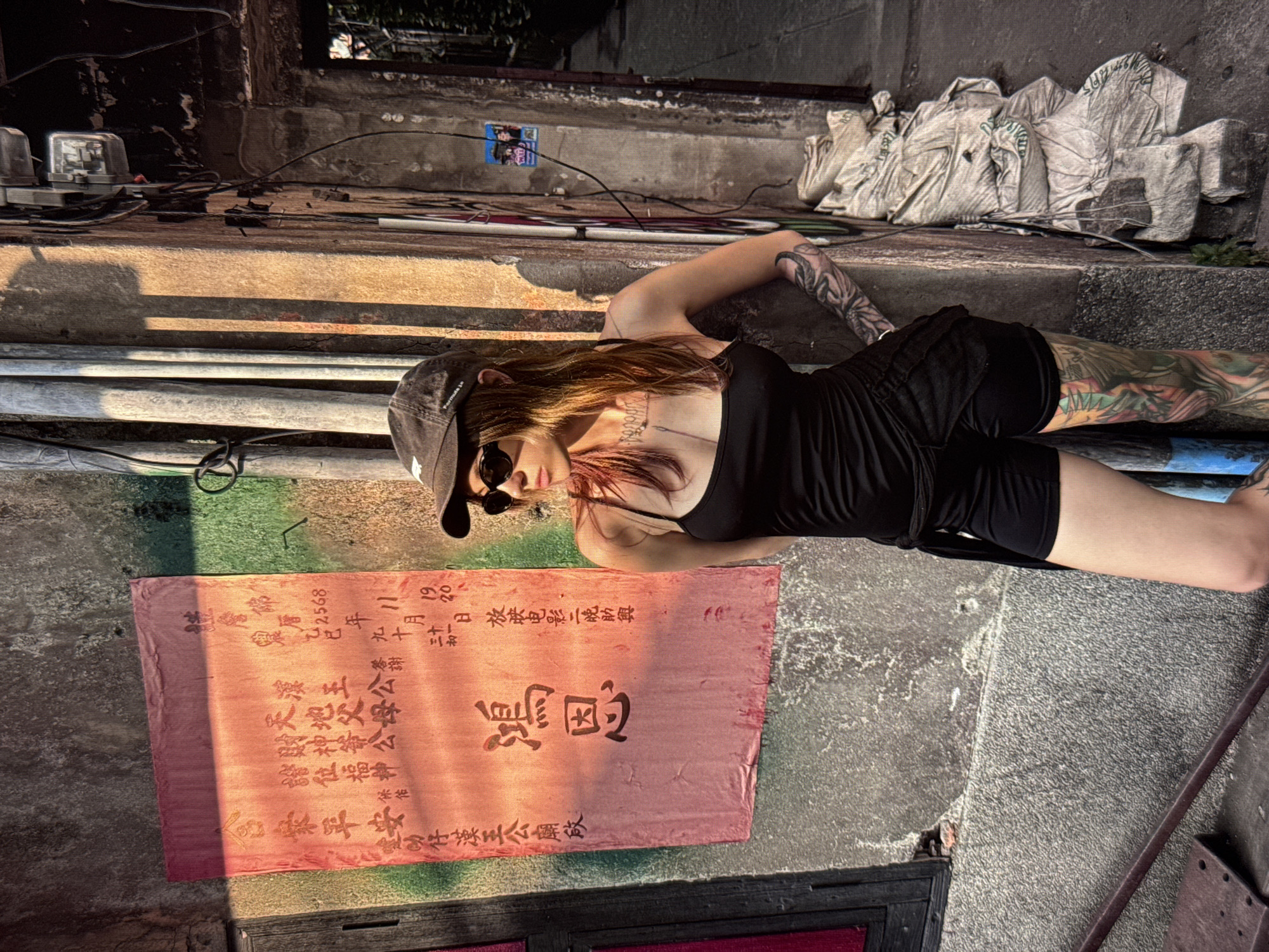 The image shows a woman with tattoos on her arms and legs, wearing a black tank top, black shorts, sunglasses, and a baseball cap. She is standing next to a wall with a red banner that has Chinese characters on it. The wall appears to be made of concrete and is weathered. There are pipes and electrical conduits attached to the wall, and a pile of what appears to be sacks is stacked against it. The scene suggests an urban environment, possibly an alleyway or a side street. The lighting indicates it is daytime, with sunlight casting shadows. There is no indication that this location is in Moscow, Russia, as there are no recognizable landmarks or cultural references to suggest that specific location.