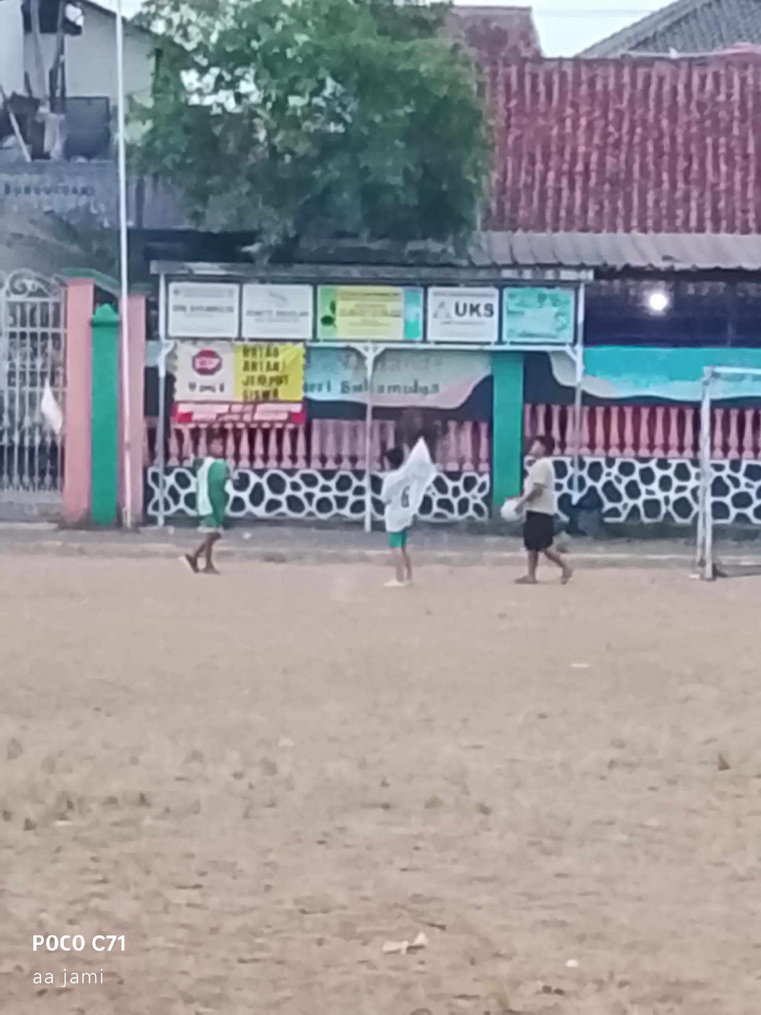 The image depicts an outdoor scene, likely a schoolyard or sports field, with a group of children playing. The ground is dusty brown, suggesting a dry, unpaved surface. In the background, there's a building with a red, corrugated metal roof, and a white and green fence. Several signs are visible on the building, one displaying "UKS", which is an abbreviation for "Unit Kesehatan Sekolah" (School Health Unit) in Indonesia.
Three children are the main subjects of the image. On the left, a child in a green and white outfit is kicking or dribbling a ball. In the center, another child, also in green and white, is holding a white flag. To the right, a third child, wearing a light-colored t-shirt and dark shorts, is holding a white ball. The children appear to be engaged in a game of some sort.
The overall atmosphere is casual and playful. The lighting suggests it's daytime, possibly late afternoon or early evening, given the soft light. The presence of educational and health-related signage points towards an educational institution. The image's metadata indicates it was taken in Tasikmalaya, Indonesia, on the POCO C71 phone by "aa jami". The image's quality is somewhat blurry, which could be due to the camera's focus, movement during capture, or the device's lens.