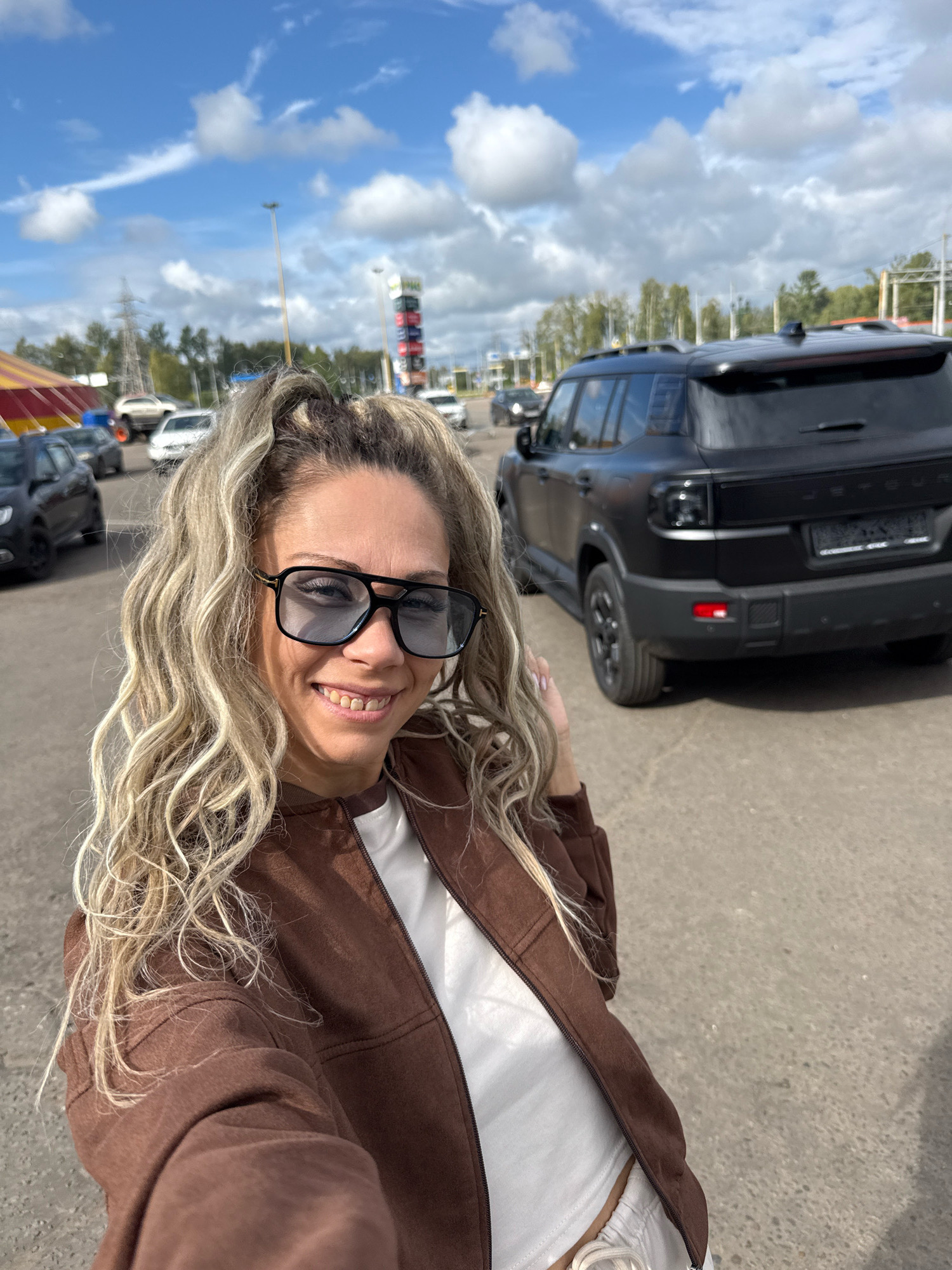 A medium shot captures a smiling woman with blonde, curly hair, wearing large sunglasses, a brown jacket, and a white top. She is standing outdoors on a paved surface, with a black SUV parked behind her. The background shows a cloudy blue sky, trees, and what appears to be a tent with yellow and red stripes, possibly part of a circus or outdoor event. The location is identified as Rusanovo, Russia. Several other vehicles are visible in the distance.
