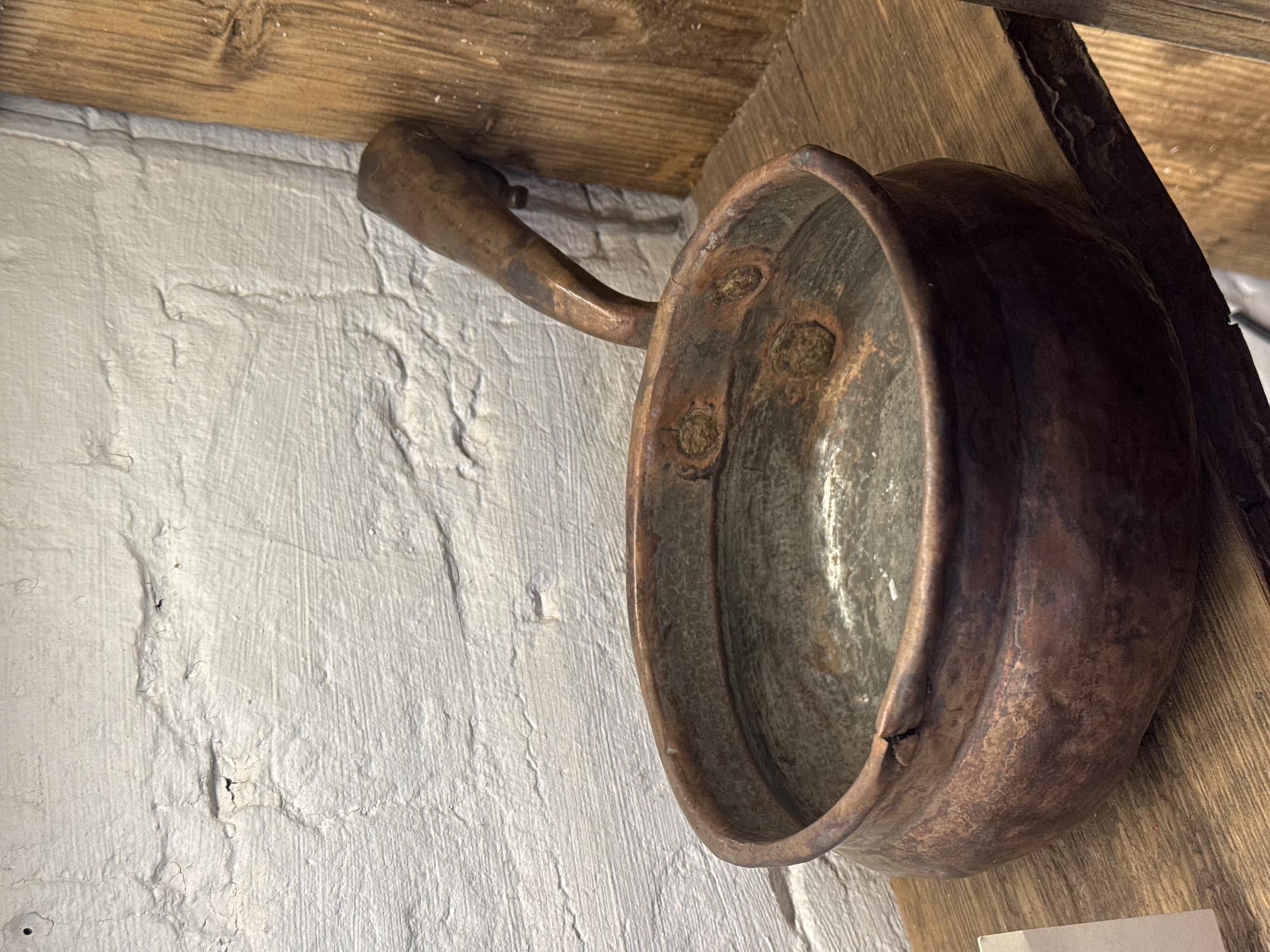 An antique copper cooking pot with a curved handle hangs on a wall. The pot is tarnished, with visible corrosion and dark spots. Three circular indentations with material inside are visible on its side. The pot is positioned against a textured white wall and a weathered wooden beam. Another wooden beam is visible on the right, with a dark metal object leaning against it. The scene is indoors, with a rustic aesthetic. There is no visible evidence indicating the city of Novo-Nikol'skoe, Russia, in the image.