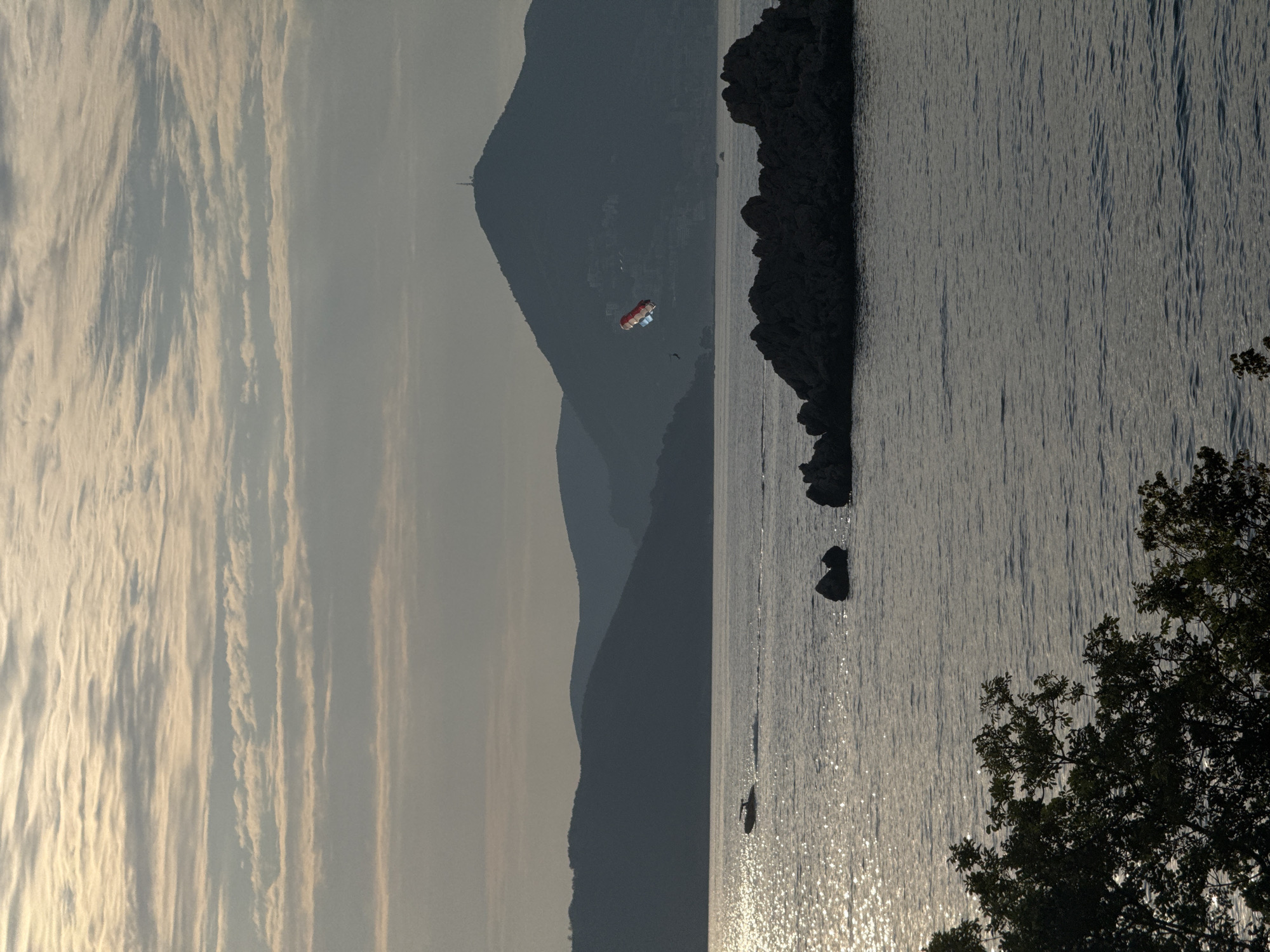This media file captures a serene waterscape with a backdrop of layered mountains and a silhouetted coastline. The scene is dominated by the calm, shimmering surface of the water, reflecting the soft, diffused light of what appears to be either early morning or late afternoon. Wispy, elongated clouds stretch across the sky, adding a textural element to the upper portion of the frame.
In the mid-ground, a parasailer is visible, suspended mid-air. The parasail is red, white, and blue, a vibrant spot against the muted tones of the mountains behind. The mountains themselves are dark and imposing, with a softer, haze-like appearance in the distance, suggesting depth and scale. Along the rocky coastline, dark, rugged formations rise from the water.
Further out on the water, two small boats are visible, one appearing to be a kayak or paddleboarder, and another possibly a small motorboat. The sunlight glints off the water's surface, creating a dazzling effect. In the foreground on the right, the dark silhouette of dense foliage, likely trees, partially obscures the view, adding a sense of depth and framing the scene.
The overall mood is peaceful and tranquil, with a hint of adventurous activity from the parasailer. The natural beauty of the mountainous coastline and the sea are the primary subjects, with the human elements adding a touch of dynamic contrast. The lighting suggests a calm day, likely with mild weather. While no text is visible, the scene evokes a sense of leisure and natural beauty. Given the context, this is likely Sveti Stefan, Montenegro, known for its picturesque coastal scenery.