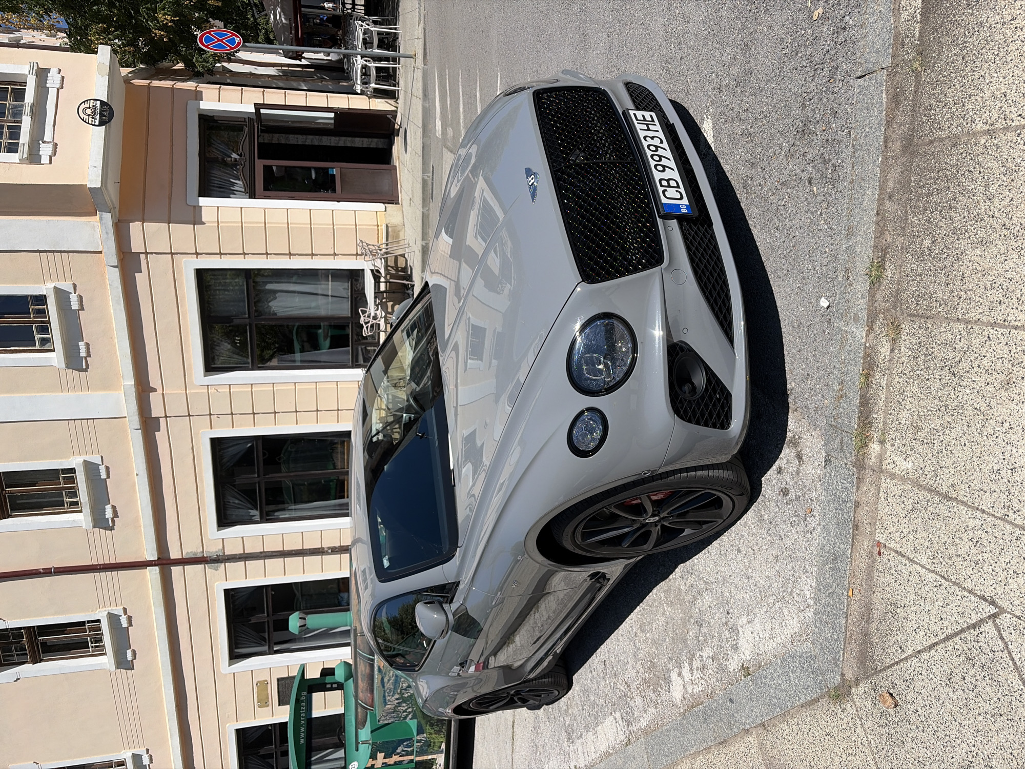 A light gray Bentley Continental GT is parked on a sunlit street in Vratsa, Bulgaria. The car is the primary subject, positioned in the foreground and occupying the center of the frame. Its distinctive grille, headlights, and license plate are clearly visible. The license plate reads "CB 9993 HE". To the left of the Bentley, a portion of a green train-like vehicle with the text "www.vratza.bg" is visible. Behind the car, a light peach-colored building with dark-framed windows and white trim stands. An open doorway leads into the building, and outdoor seating with tables and chairs is set up on the sidewalk in front of it. A no parking sign is visible on a pole to the right of the building entrance. The ground is paved with asphalt and some sections of concrete or stone walkway. The overall impression is a street scene on a clear, sunny day in a European city.