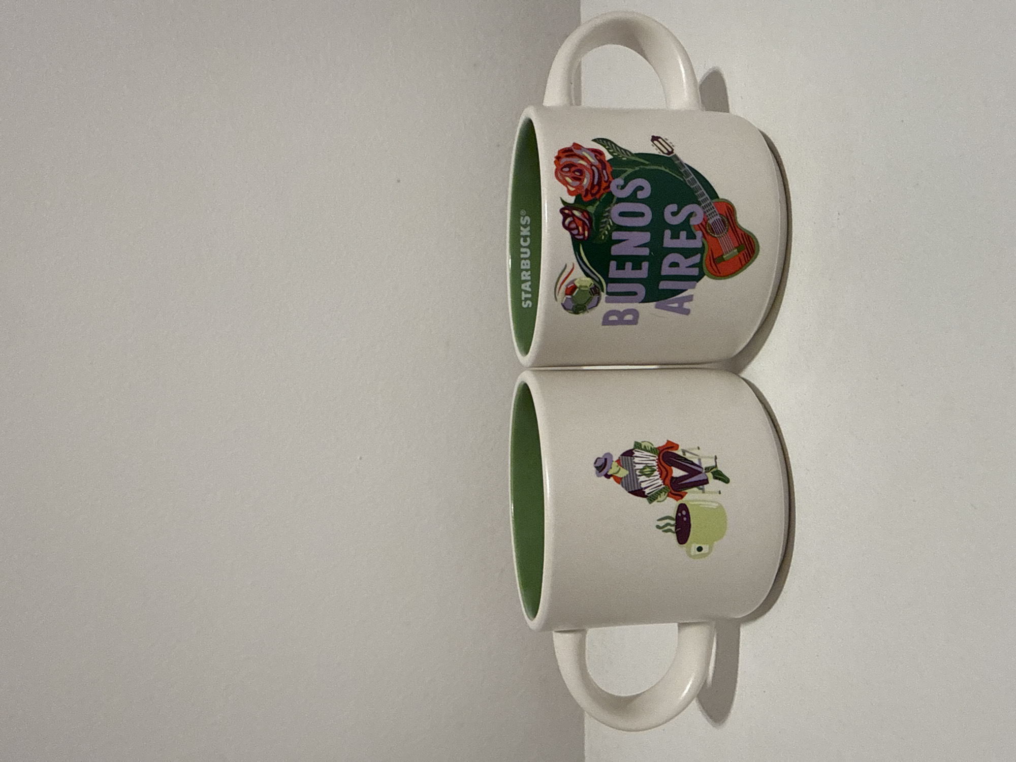 Two white Starbucks mugs, each with a bright green interior, are stacked vertically on a plain, light-colored surface. The top mug rests directly on the bottom one, and both are oriented with their handles facing left and their decorated sides visible. A shadow is cast beneath the mugs on the right side, suggesting a light source from the left.
The top mug features the word "BUENOS AIRES" prominently displayed in purple text on a green, circular background. Surrounding the text are colorful illustrations including red roses on a vine, a brown acoustic guitar, and a small, multi-colored soccer ball. The "STARBUCKS®" logo is visible in white, oriented vertically along the left edge of the mug's design.
The bottom mug has a different but related design, also on a white background. It depicts a stylized figure in a hat playing a bandoneon or accordion, which is an instrument strongly associated with tango music. To the right of this figure, there is an illustration of a green mate gourd with a bombilla (straw) and wisps of steam rising from it, representing the traditional South American drink.
No specific activity is taking place; the mugs are static. The designs on the mugs serve as direct location cues, celebrating the culture of Buenos Aires, Argentina, through iconic imagery such as roses, a guitar (tango), a soccer ball, an accordion/bandoneon, and a mate gourd. The lighting appears to be indoor, and no time of day or weather is discernible.
