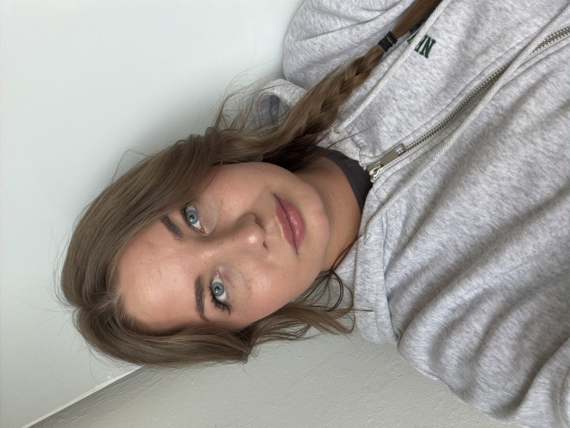 A young woman with light brown hair braided on one side and blue eyes is looking at the camera. She is wearing a light gray zip-up hoodie with dark green lettering. The background consists of a white wall and corner. There is no indication of the location being the city of Chaikovsky, Russia.