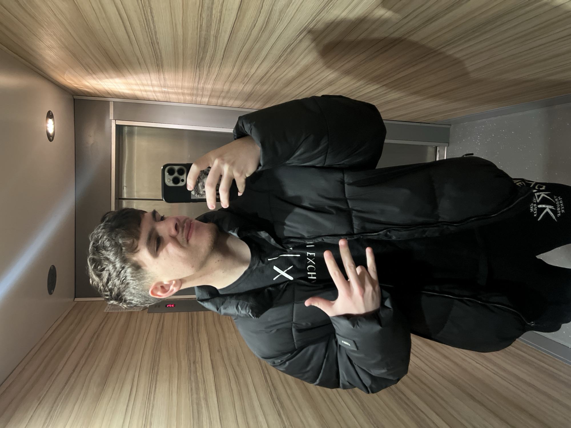 A young man is taking a selfie in what appears to be an elevator. The interior is lined with light-colored wood paneling. The elevator has a stainless steel door and a light fixture on the wall. He is wearing a black puffer jacket over a black t-shirt with white text. He is holding a smartphone in his left hand to capture the image, while his right hand is making a hand gesture with his fingers extended. There is no indication of the city of Sofia, Bulgaria, in the image.