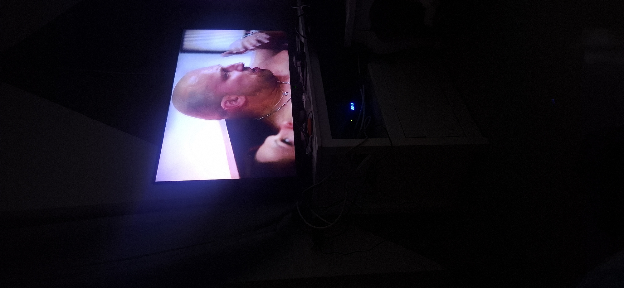 In a dimly lit room, a large flat-screen television displays a scene featuring two individuals. The primary focus on the screen is a bald, fair-skinned man with a muscular build, wearing a necklace. He is looking towards his right and appears to be speaking, with his hand raised in a gesture. To his left, a woman with dark hair is partially visible, looking towards the man. The television is positioned on a white cabinet, on which several electronic devices and cables are present. One device on the cabinet displays a blue digital clock reading "23:13". The overall environment suggests a domestic setting at night. The image is a photograph taken in Yerocham, Israel.