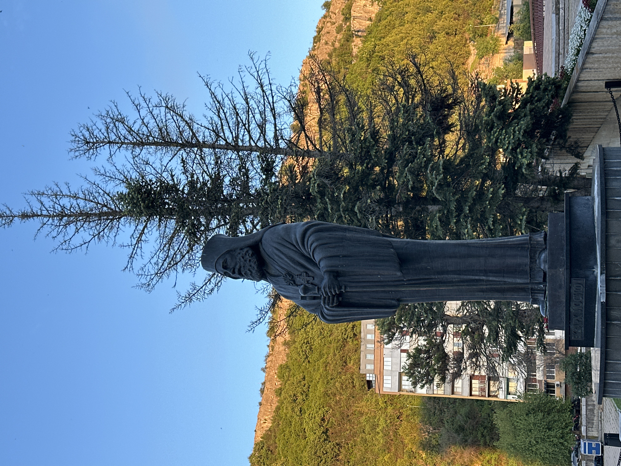 A vertical, eye-level shot shows a statue of a cleric holding a cane in his right hand, set against a backdrop of lush greenery and a building. The statue is dark, with a weathered appearance. Behind the statue are trees, some of which appear dead or bare. The background features a steep, rocky cliff covered in green vegetation. In the distance, a building with multiple windows is visible. The sky is clear blue. The inscription "EXARCH JOSEPH I" can be seen at the base of the statue. The location is likely Lovech, Bulgaria, given the statue's prominent presence there.