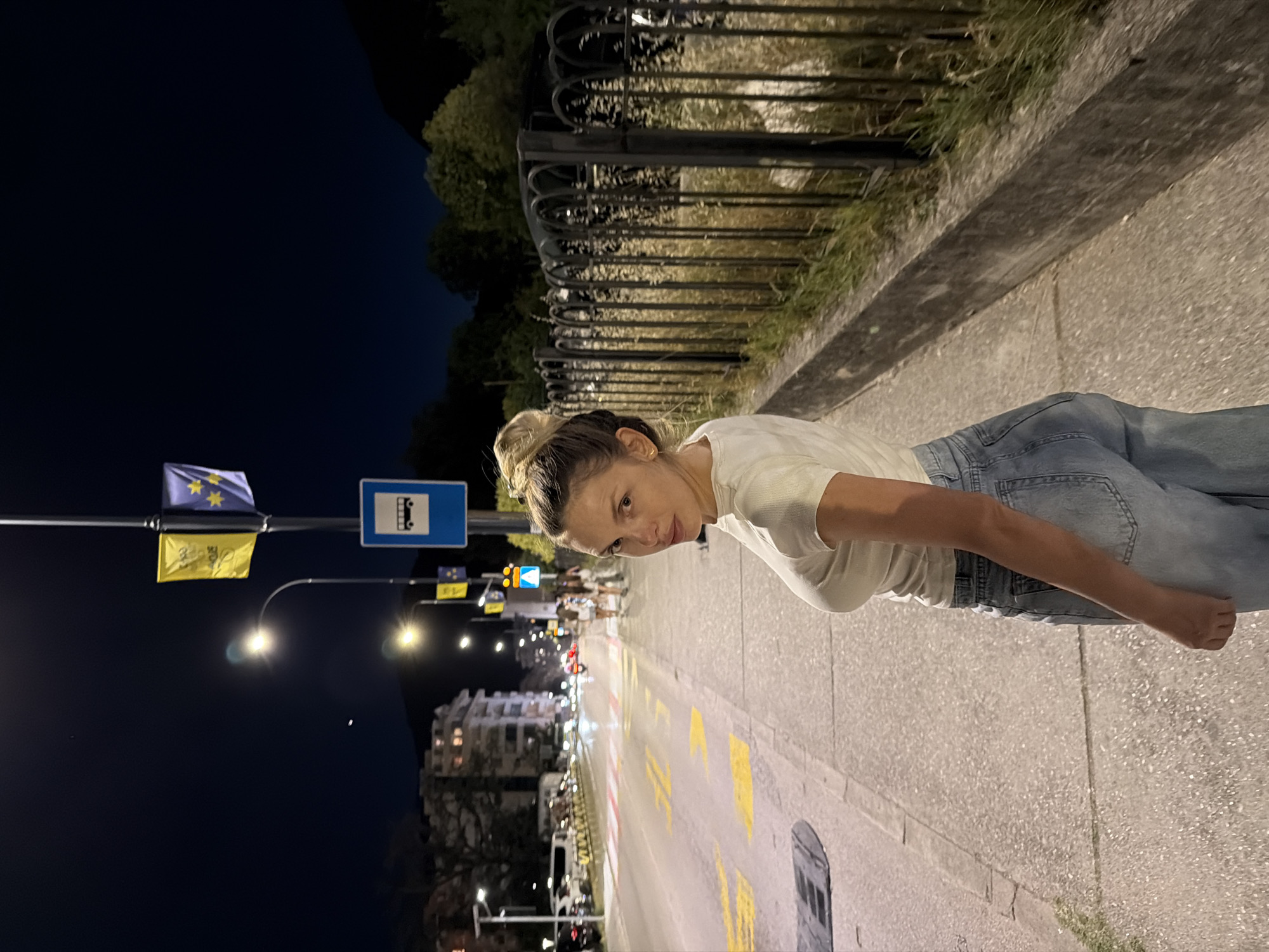 A young woman with blonde hair in a bun, wearing a white t-shirt and light blue jeans, stands on a sidewalk at night. She is looking back over her right shoulder at the camera. The sidewalk is made of textured concrete and is illuminated by streetlights. To her left, a street runs past with cars, a crosswalk with red and white markings, and yellow directional arrows painted on the asphalt. Buildings are visible in the background on the left side of the street, with illuminated windows. To her right, a black metal fence with decorative arches runs along the edge of the sidewalk, with dry grass and some greenery growing behind it. Overhead, a street lamp with a curved arm illuminates the scene, and a blue bus stop sign with a white bus icon is visible. Two flags, one yellow and one blue with yellow stars, are also attached to a lamppost above the bus sign. The sky is dark blue. The location is identified as Budva, Montenegro.