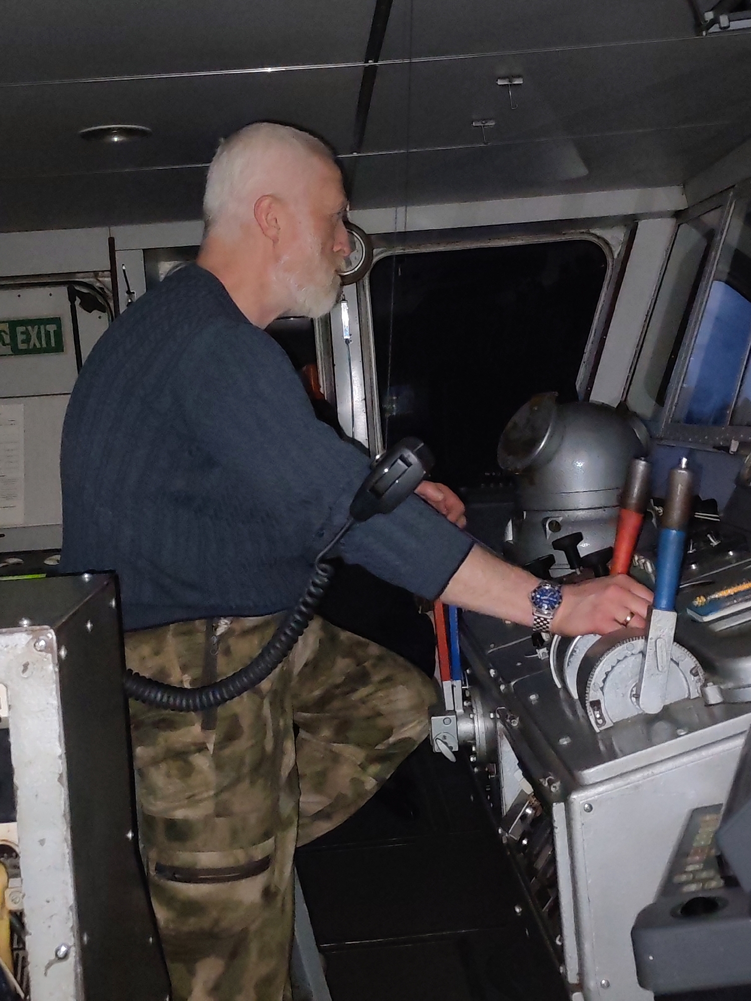 A man stands in what appears to be the control room of a ship or vessel, operating a control panel. He is Caucasian, with short white hair and a white beard. He wears a dark blue sweater and camouflage patterned pants. His left leg is partially visible, appearing to be elevated. He is holding a microphone in his left hand and manipulating a lever on the control panel with his right. He is wearing a watch on his left wrist. The control panel is metal and features multiple levers and controls. Behind the man is a window showing a dark view of the outside. An "EXIT" sign is visible on a wall to the left. The location cannot be determined as being in the city of Uzhla, Russia based on the provided visual information.