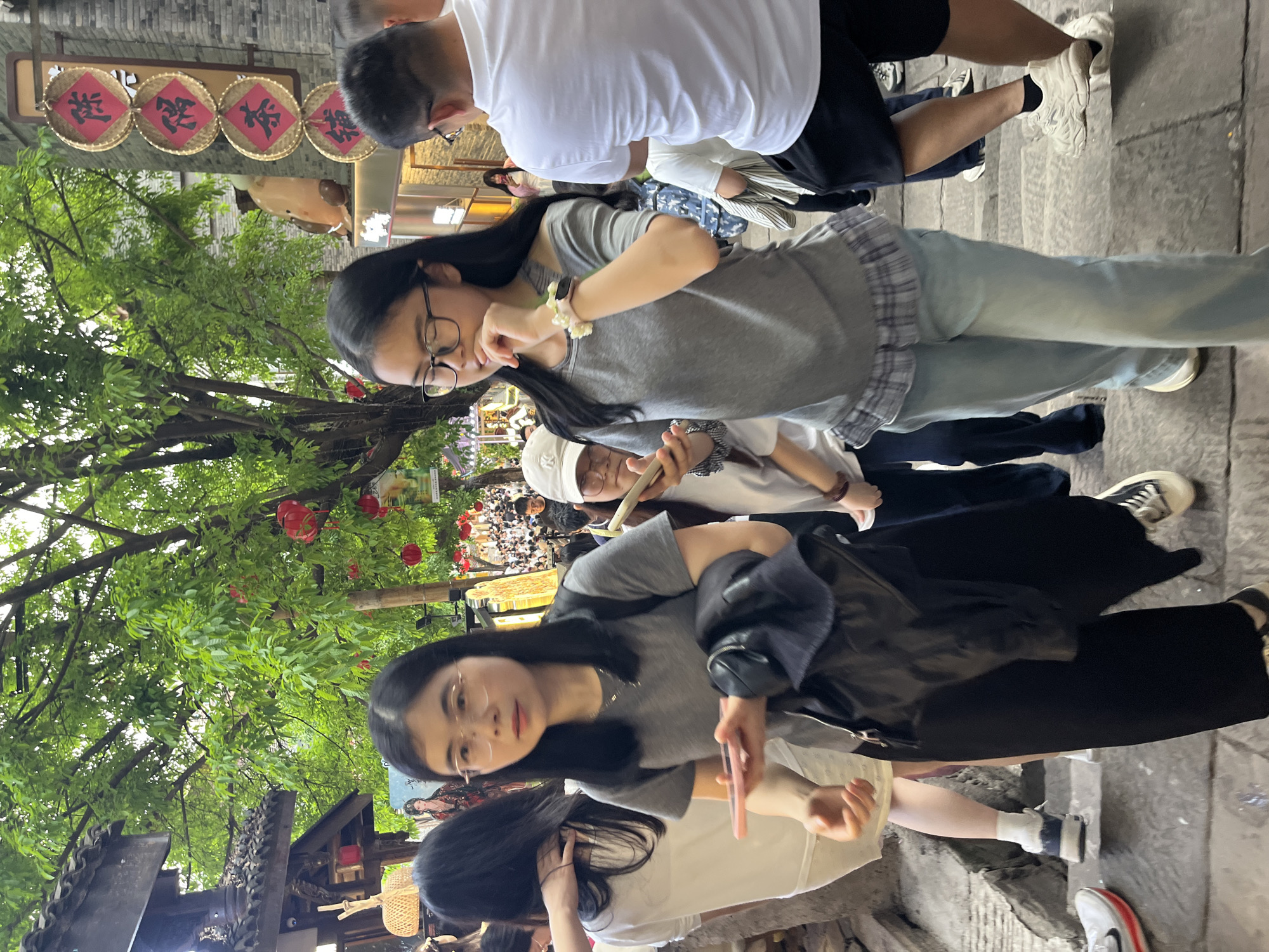 The image depicts a bustling outdoor scene in what appears to be a pedestrian street or cultural district, consistent with Yuzhong District, China. The perspective is slightly elevated, capturing a crowd of people amidst traditional architecture and lush greenery.
In the foreground, two young women are prominent. The woman on the left, with long dark hair and eyeglasses, wears a grey t-shirt and black trousers, holding a pink smartphone and looking towards the camera. The woman on the right, also with long dark hair and glasses, wears a grey t-shirt, light blue jeans, and has a plaid shirt tied around her waist; she is partially obscured by another person but also appears to be engaging with the camera with a slight hand gesture.
Various other individuals are visible throughout the frame, including a man in a white t-shirt and dark shorts (seen from behind) and another person wearing a white cap holding a thin object. The setting features significant green foliage from overhead trees, from which several red spherical lanterns hang, adding a festive touch. Traditional dark-tiled roofs and intricate architectural details are visible on the left side. The ground is paved with stone.
In the upper background, four circular, woven signs are displayed on a building facade, each featuring a single red Chinese character. From left to right, these characters are "送" (sòng), "祝福" (zhùfú), "吉祥" (jíxiáng), and "如意" (rúyì), which collectively translate to "Send blessings, auspiciousness, and good fortune." The lighting suggests daytime, possibly an overcast day, providing soft, diffuse light. The scene conveys a lively and culturally rich atmosphere.