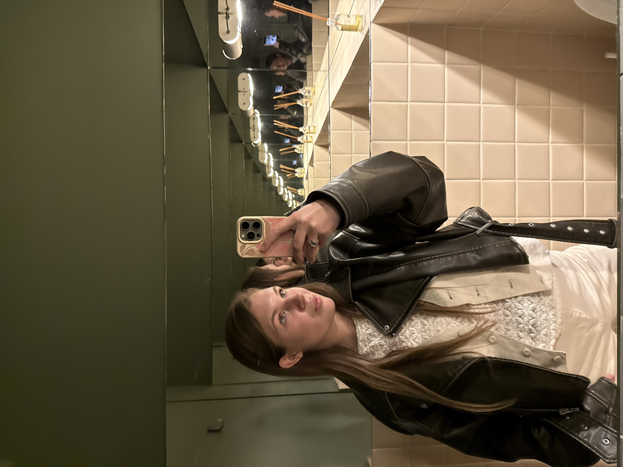 A young woman is taking a selfie in a bathroom with multiple mirrors, creating an infinity mirror effect. She is holding a smartphone with a visible triple-lens camera in her right hand. She is wearing a dark leather jacket over a white top. The bathroom has light-colored square tiles on one wall and a dark green wall on the other. There are white cylindrical light fixtures above and a diffuser with reed sticks reflected in the mirrors. The location appears to be in Belgrade, Serbia.