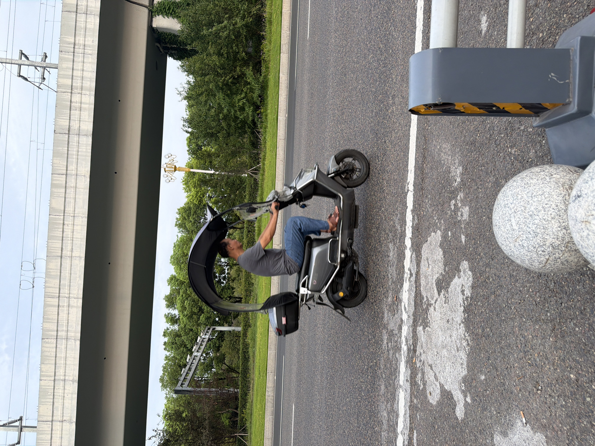 A man rides a grey electric scooter equipped with a dark fabric canopy on an asphalt road. The scooter has a black storage box mounted behind the seat. The man wears a grey t-shirt and blue jeans. The road is lined with a white stripe. In the background, there is a green lawn and trees. Above the trees, a concrete overpass runs horizontally across the image, with electrical wires suspended from poles. A yellow street lamp is visible to the left of the overpass. In the foreground, a grey concrete bollard with yellow and black hazard stripes is partially visible on the right side of the image, and two round, speckled stone bollards are on the left. The scene appears to be in the city of Jiangjin, China.