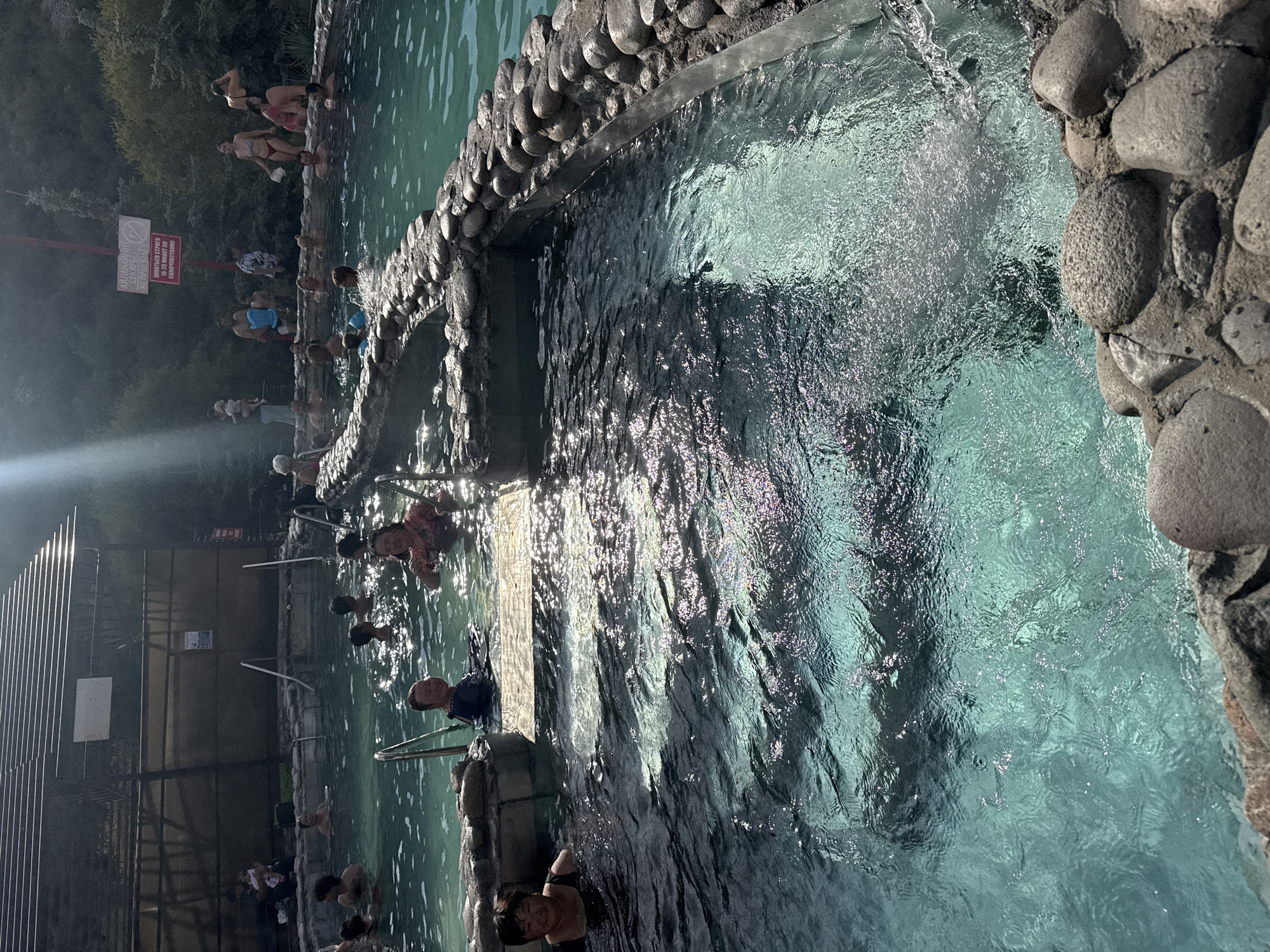 The image shows an outdoor hot spring in Ak-Suu, Kyrgyzstan. Several people, including adults and children, are bathing in the turquoise water. The hot spring is surrounded by a stone border and a wooden structure is visible in the background. Sunlight is streaming down, creating a shimmering effect on the water's surface. Some individuals are sitting on the edge of the pool, while others are submerged in the water. There are also signs posted in the background, suggesting a recreational area.