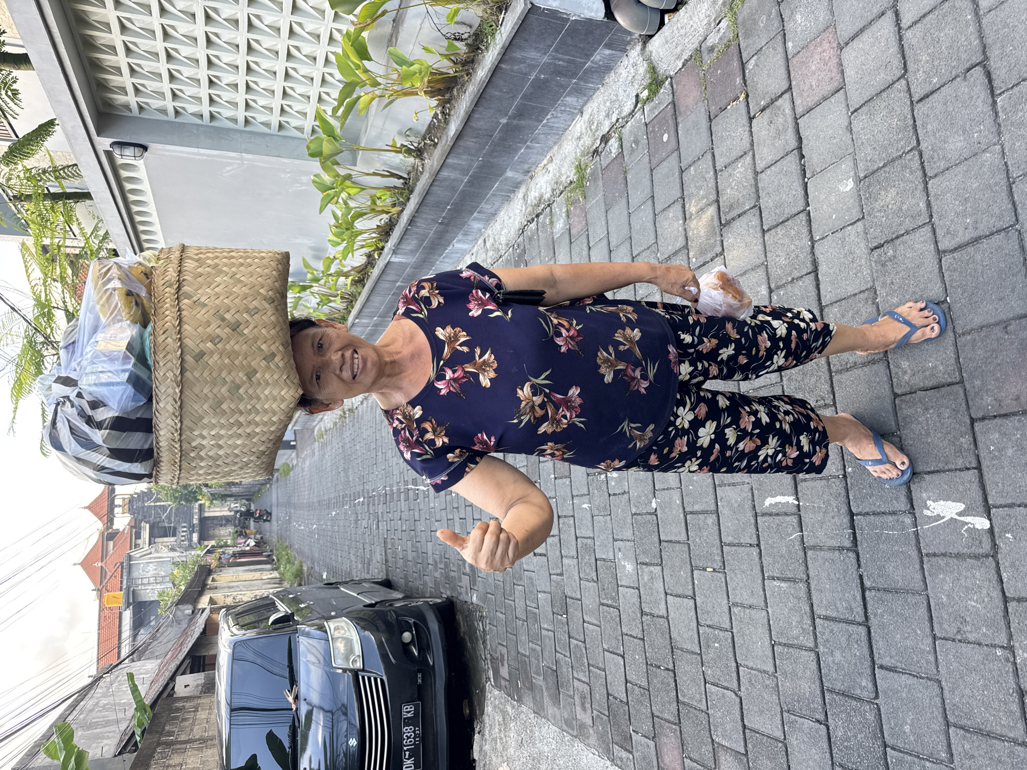 A woman stands on a brick-paved street in Kuta, Indonesia. She balances a large woven basket on her head, which contains a variety of items wrapped in plastic. She wears a blue floral shirt and dark pants with a similar floral pattern and blue flip-flops. In her right hand, she holds a plastic bag containing food. The woman is smiling and giving a thumbs-up gesture. Behind her, a black van is parked, with a license plate partially visible, reading "DK 1638 KB." The street leads into a residential area with houses and greenery visible. The architecture appears to be local Indonesian style.