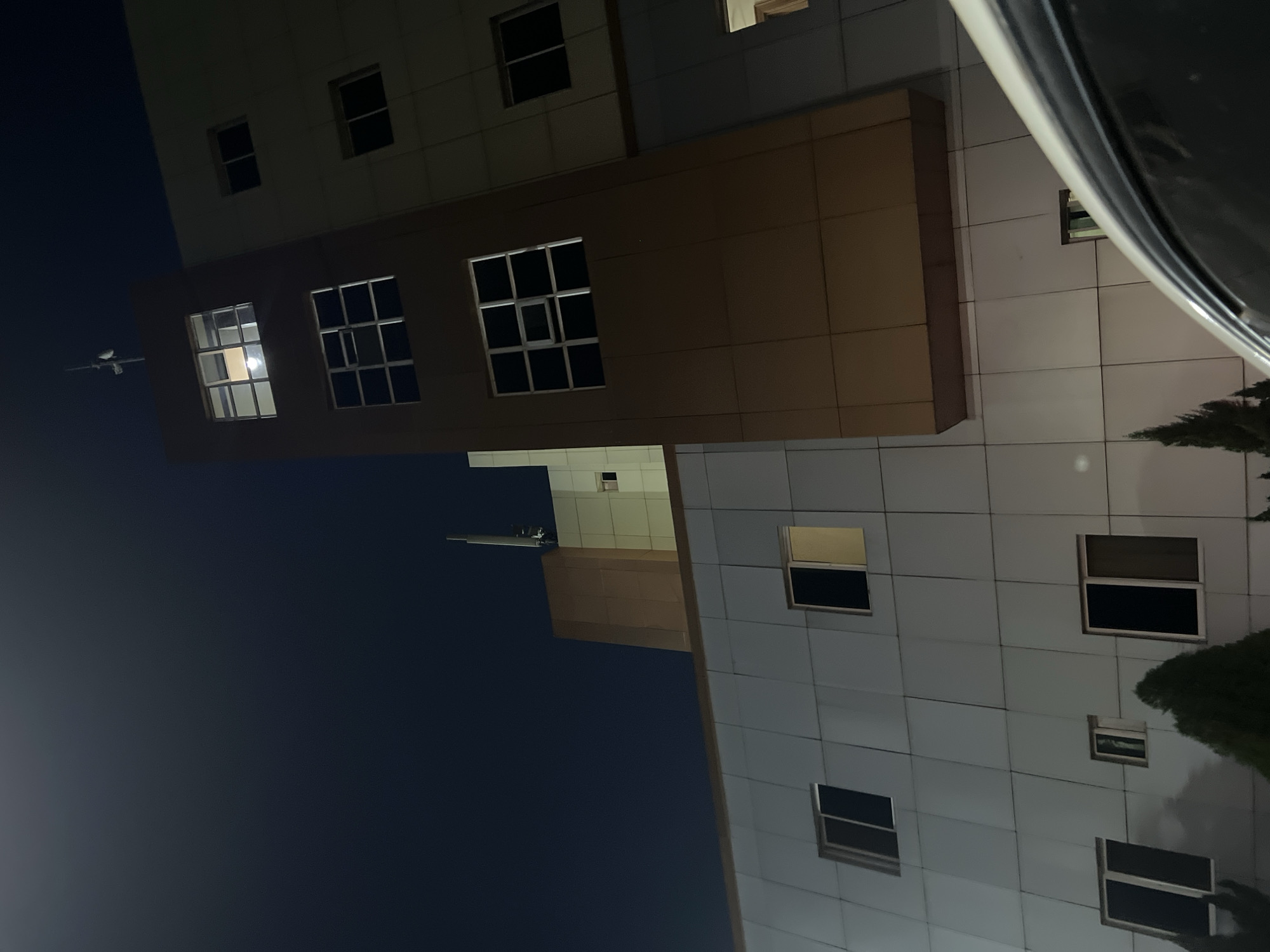 An outdoor, eye-level shot captures modern buildings against a dark night sky in Abuja, Nigeria. The image is oriented vertically but depicts buildings typically seen horizontally. On the left, a multi-story building features sections with a light brown, tiled facade and several rectangular windows. One window on this brown section is brightly lit from within. Above this, another section of the building, possibly beige or light grey, also has dark windows.
In the upper left portion of the frame (when correctly oriented), a small, white aircraft—likely a plane or drone—is visible in flight against the deep blue-black sky.
To the right, another large building with a light-colored (possibly white or light grey) paneled facade dominates the frame. This building also features numerous rectangular windows, some dark and some showing faint light or reflections. Dark vegetation, possibly trees or shrubs, can be seen along the lower right edge of this building. The reflective surfaces of this building create subtle light patterns.
In the top right corner, a dark, curved object, possibly part of a vehicle's roof or another foreground structure, intrudes into the frame. The scene is illuminated by internal building lights and ambient light, with no visible text. The overall impression is an urban nightscape.