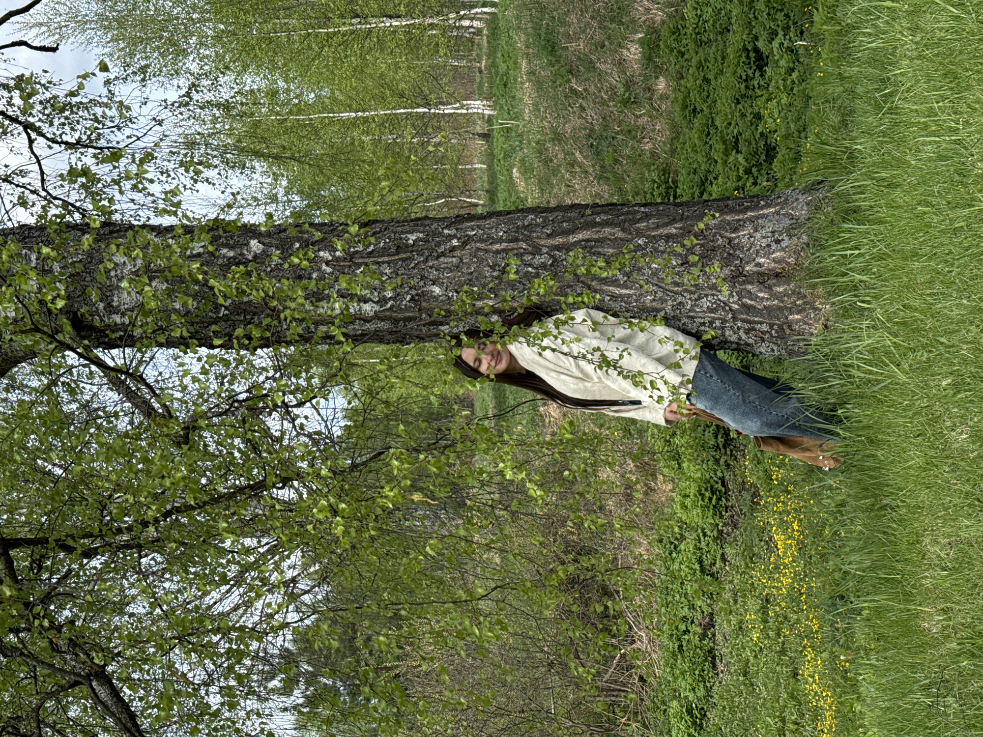 A young woman leans against a tree trunk in a grassy area. The location appears to be a forested region within Красный Маяк, Russia, although specific landmarks are not discernable. The woman is wearing a light-colored shirt and jeans. She is facing the viewer and appears to be smiling. The background is a mix of trees and grass.